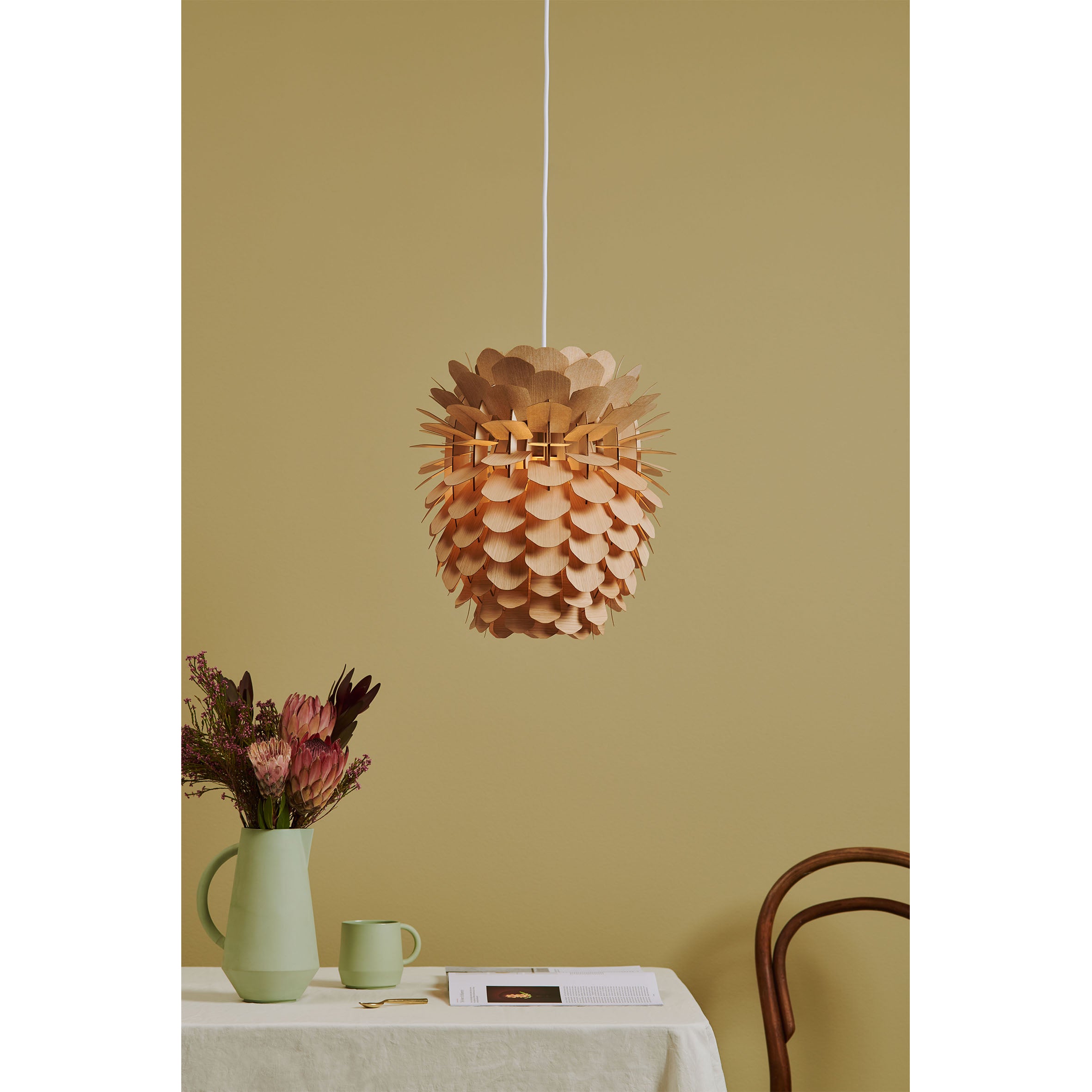 The Schneid ZAPPY ASH pendant light, crafted from ash veneer and resembling a pine cone, hangs from the ceiling. Below, a table with a beige cloth features a green jug of flowers, a matching mug, and a book. A wooden chair blends with the muted yellow wall.