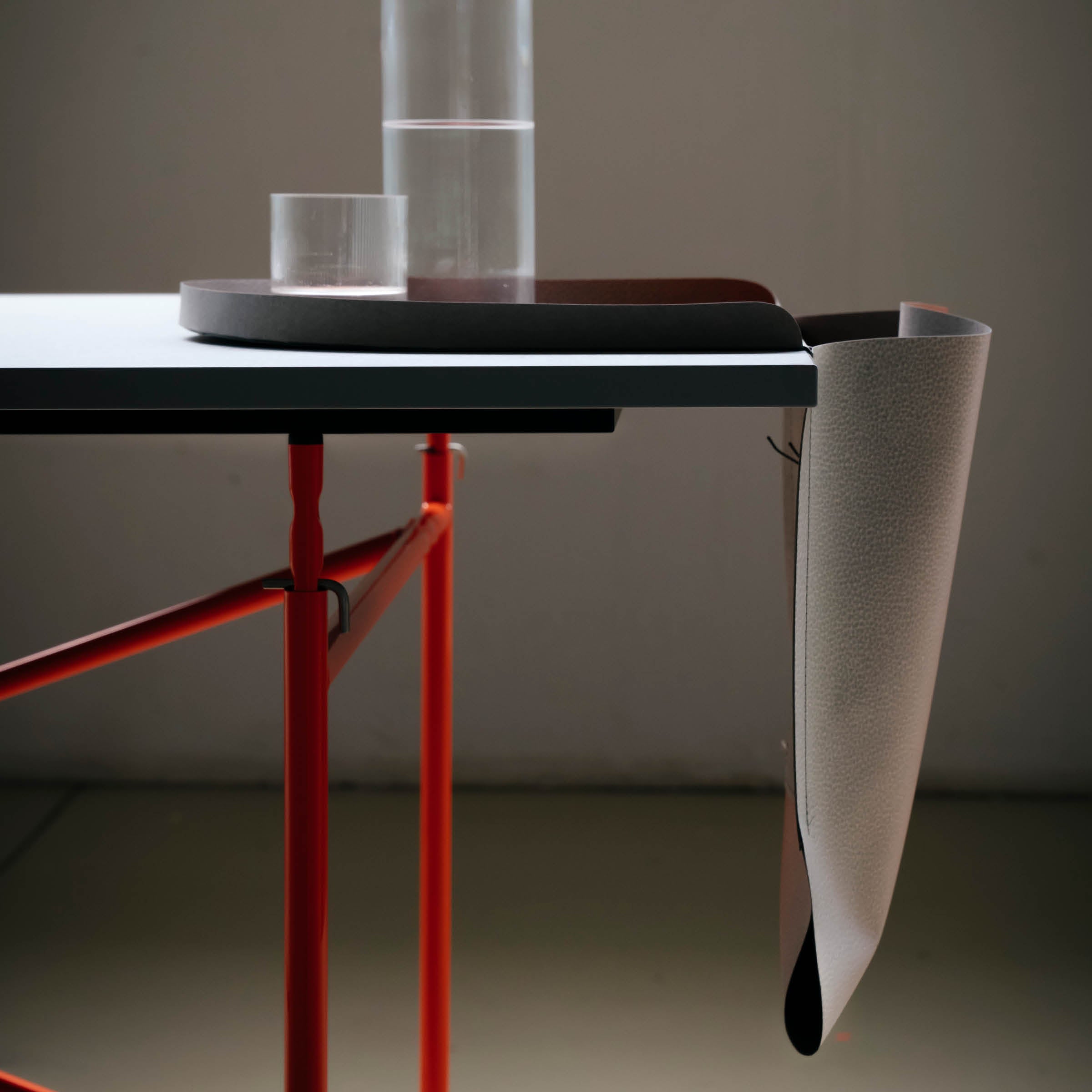 A minimalistic setup features Richard Lamperts sleek table with a white surface and red legs, crafted sustainably. The Salto Tray supports two glass containers, while a folded beige Papyr® cloth drapes over the edge. Soft lighting highlights this German-made elegance.