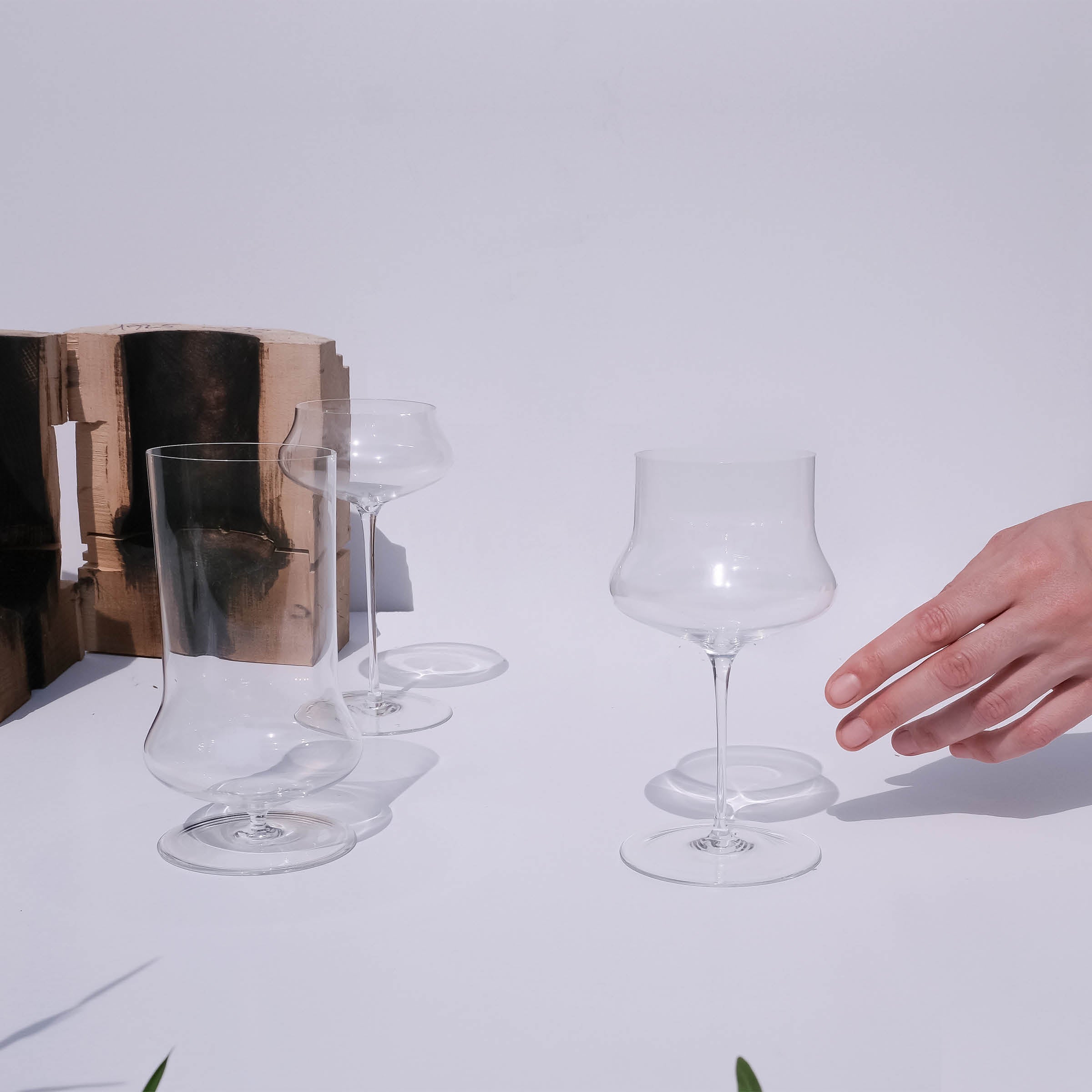 A hand reaches towards three uniquely-shaped, empty glasses from Lobmeyrs Drinking set no.287 Ident, crafted from lead-free crystal and placed on a white surface, with a wooden block casting soft shadows in the background under bright lighting.