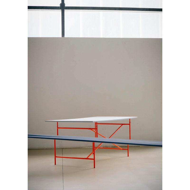 Positioned against a plain wall is a modern table with a white top and bright red metal legs, boasting an angular, geometric design perfect for contemporary settings. In the foreground, the blurred edge of the OneTwo Mirror by Richard Lampert subtly enters the frame.