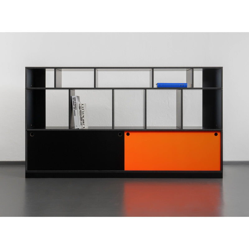 The Egal Highboard by Moormann features open compartments and sleek sliding doors. A contemporary steel frame complements the left black door, contrasting with the vibrant orange on the right. It showcases shelves with books and a blue rectangular object against a plain white wall and gray floor.