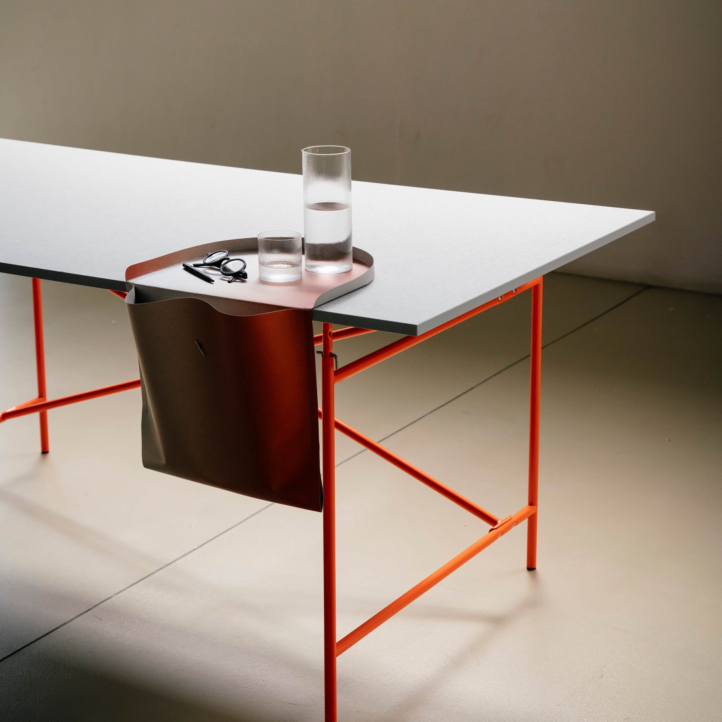 The minimalist table features an orange metal frame with a flat white Papyr® surface and a brown side pocket. It holds the Salto Tray by Richard Lampert, topped with a glass water carafe, a small glass, and scissors—proudly made in Germany.