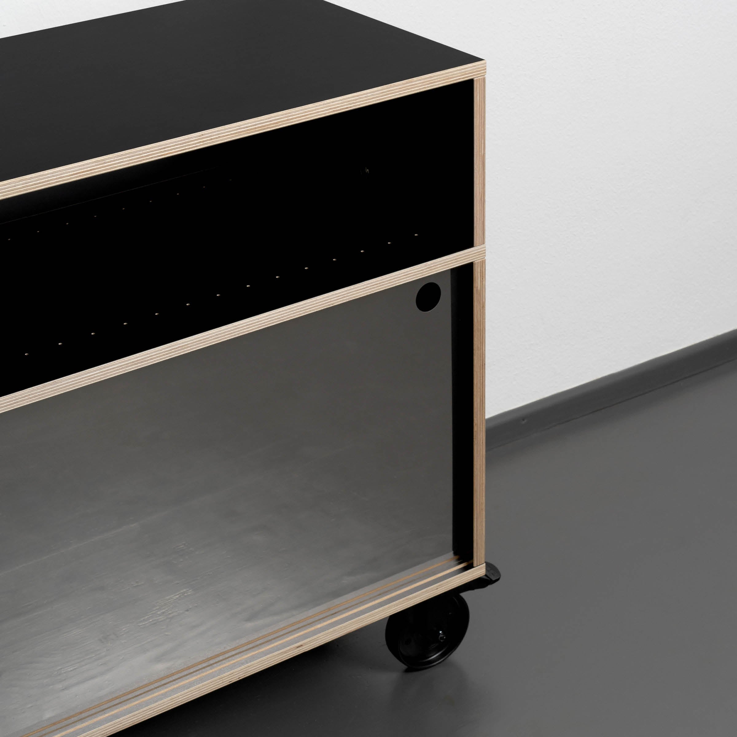 The Moormann Egal Rockboard is a modern wooden cart with a black top and open lower shelf, featuring two black wheels on one side. Its sleek design stands elegantly on a dark floor against a white wall, resembling a stylish sideboard.