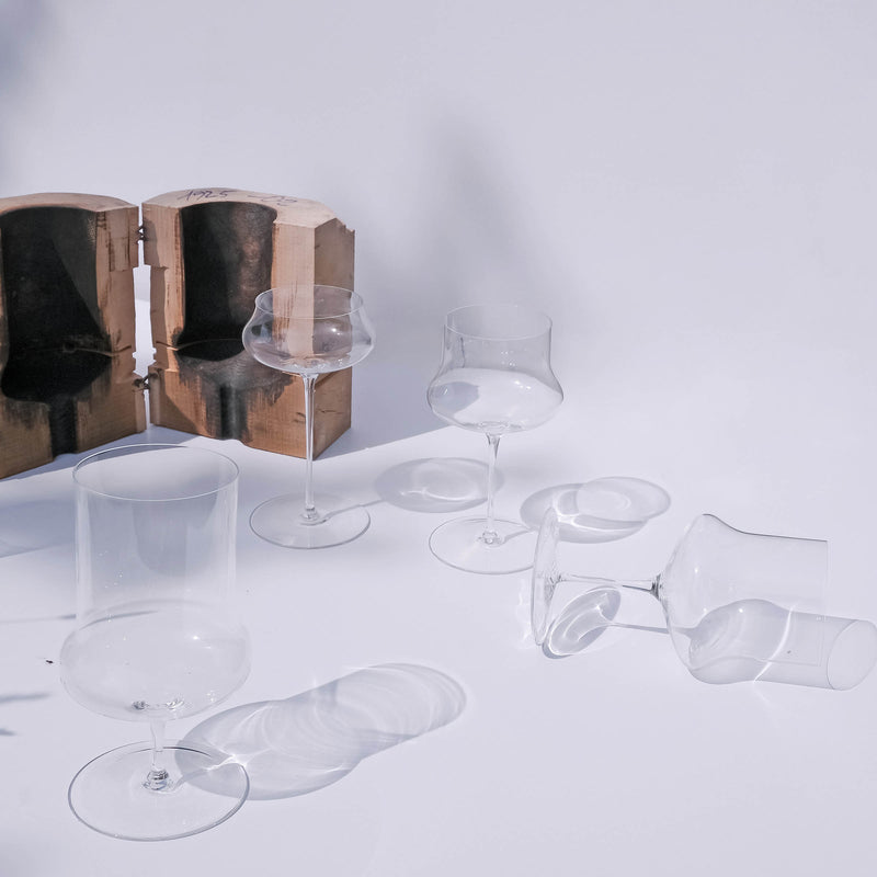 The Lobmeyr Drinking set no.287 Ident features clear, uniquely shaped wine glasses made from mouth-blown glass arranged on a white surface with soft shadows. Wooden molds by mischertraxler studio in the top left underscore the glassblowing process, creating a minimalistic and bright scene.