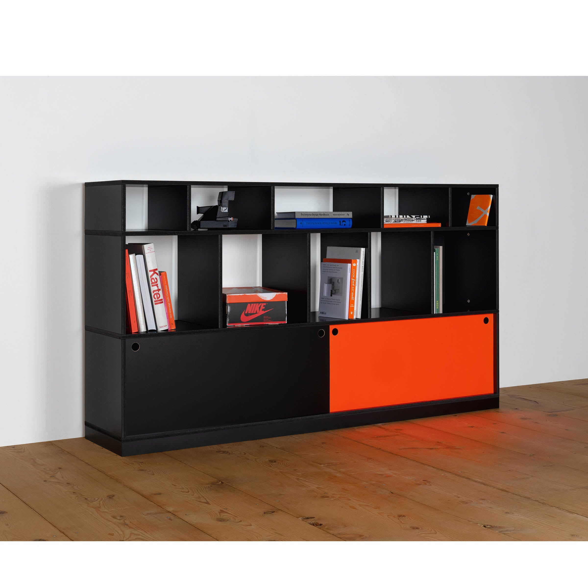 The Moormann Egal Highboard is a contemporary shelving unit in black, featuring both open and closed compartments with sliding doors for versatility. An orange panel provides a colorful accent, perfect for displaying books and decor against a white wall on wooden flooring.