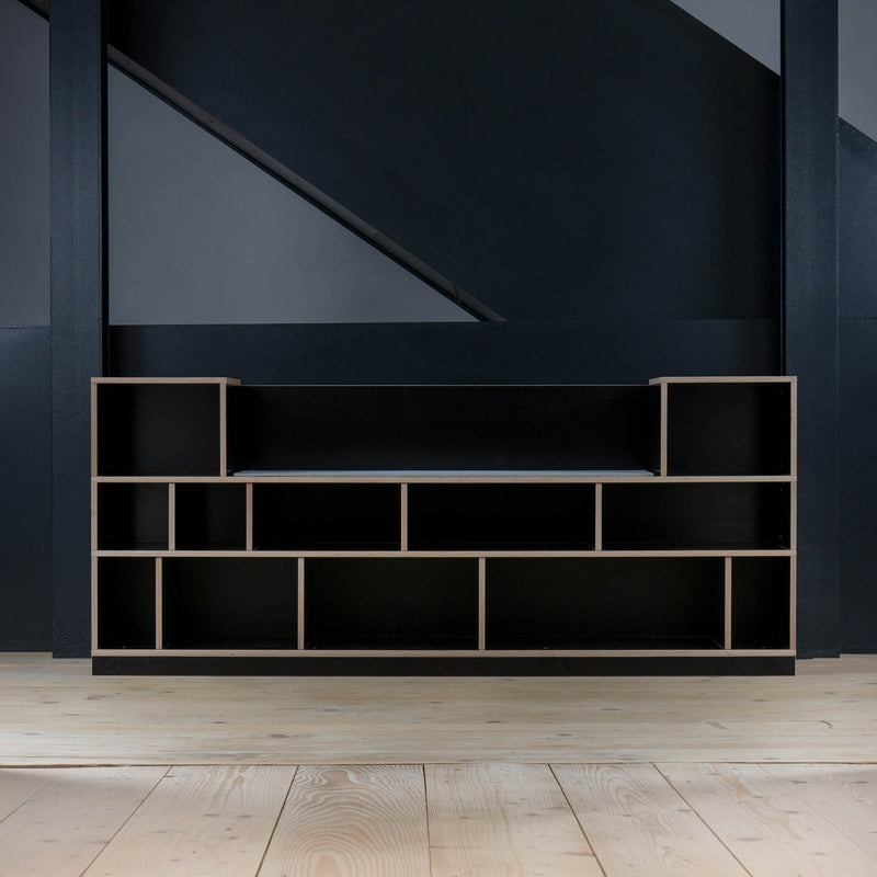 The Moormann Egal Sitzboard, a minimalist black shelving unit with open compartments and customizable sliding doors, contrasts the dark wall and complements the light wood plank flooring, enhancing the darker furniture tones.