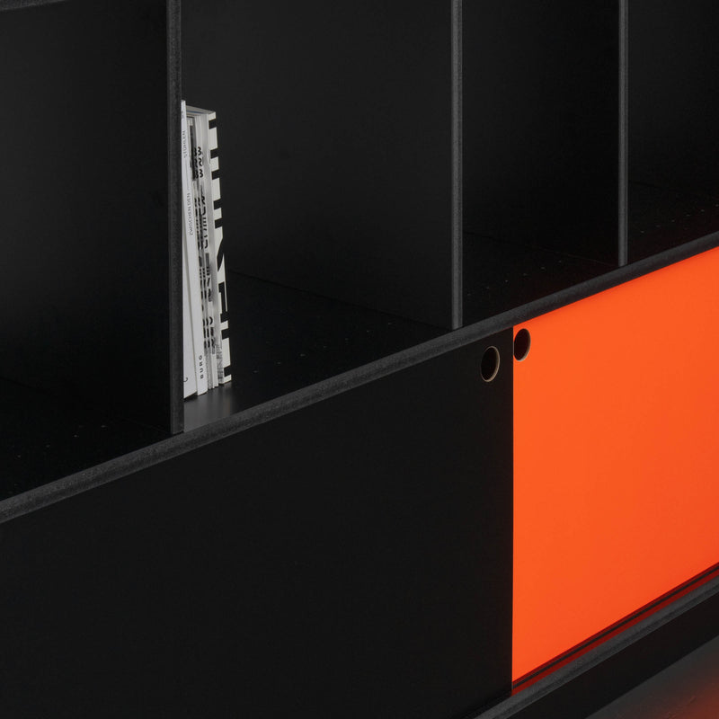 The Egal Highboard by Moormann is a black sideboard with a modern, minimalist design. It features multiple compartments, a striking orange sliding door, vertical magazine storage in one section, and sleek steel accents.