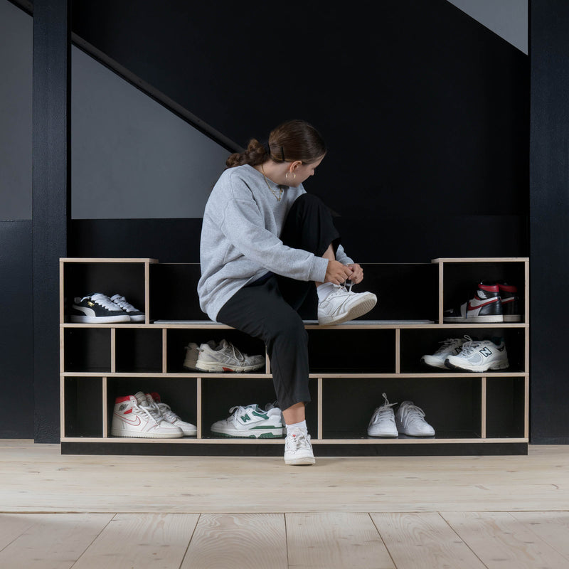 Sitting on a staircase with the Egal Sitzboard by Moormann, a person adjusts their shoe. They wear a gray sweater and black pants. The staircase includes a wooden shoe rack with customizable cubbies, neatly organizing an array of sneakers.