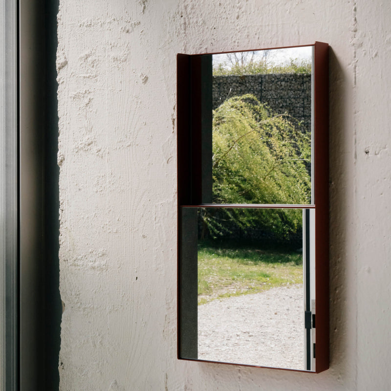 The OneTwo Mirror by Richard Lampert, a rectangular wall mirror with a minimalist metal frame, reflects greenery and a stone wall outside. Mounted on a white textured wall, it captures a partial outdoor scene through a nearby window.