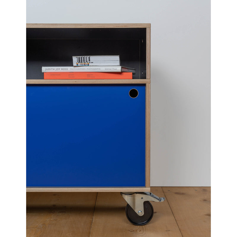 The Egal Rollboard by Moormann is a mobile storage unit with a blue sliding door, exposed plywood edges, and a top shelf for books like Judith Joy Ross. It rolls effortlessly on metal wheels against a light wood floor and offers versatile sideboard versions.