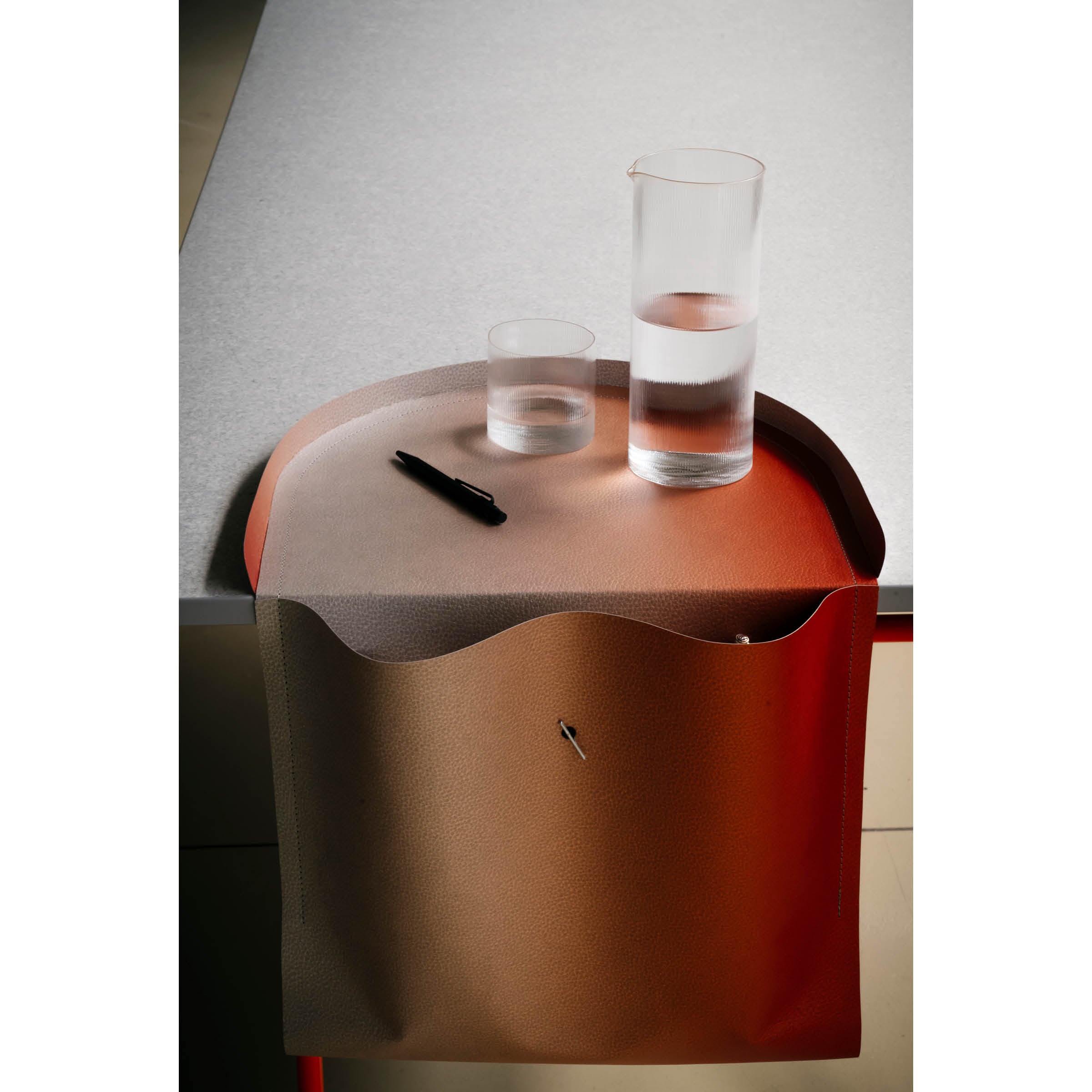 A minimalist image features the Salto Tray by Richard Lampert, a curved edge table in sustainable Papyr® material. It holds a glass of water, a cylindrical carafe, and a black pen. The side leather pocket adds modern functionality and design.
