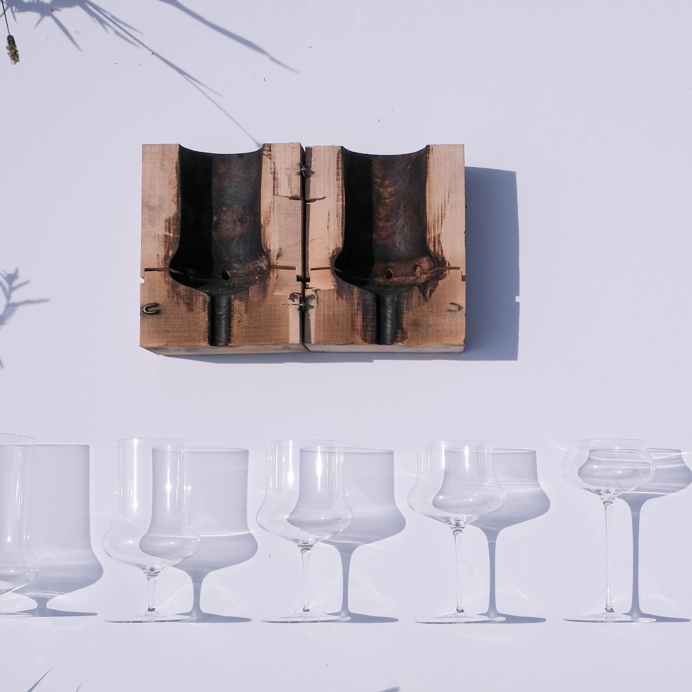 A set of six clear Ident goblets from Lobmeyr, crafted with lead-free crystal, is elegantly displayed on a white surface. Above them lies a vintage wooden mold containing two dark metal shapes, casting soft shadows for an artistic contrast.
