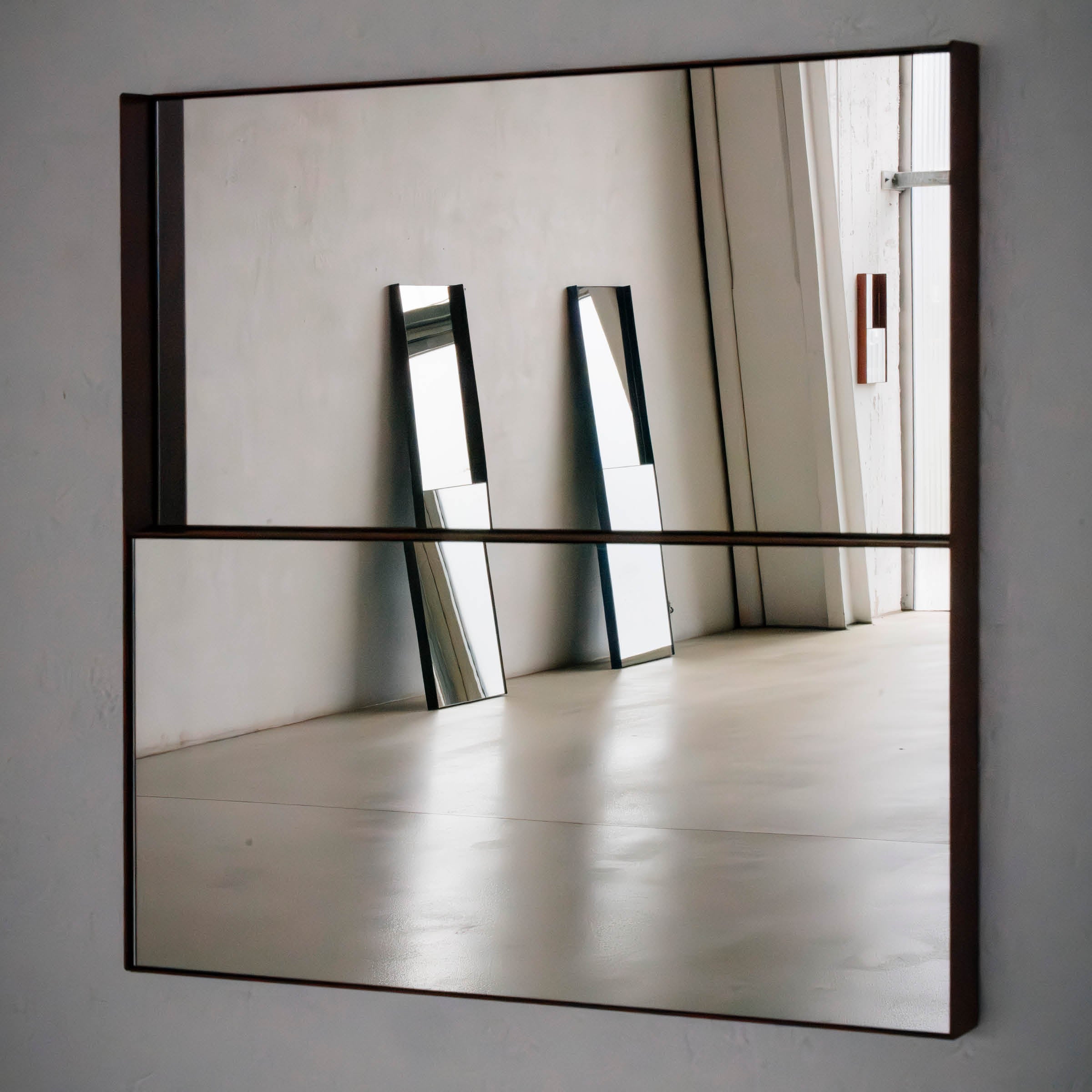 The OneTwo Mirror by Richard Lampert, with its minimalist metal frame, is mounted on the wall and reflects two tall narrow mirrors opposite. The room showcases contemporary home accessories featuring grey tones, concrete flooring, and an open doorway allowing natural light.