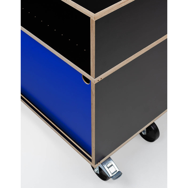 The Egal Rollboard by Moormann is a minimalist rolling cart, featuring a blue exterior with black accents. It has caster wheels for mobility, a plywood edge, and a square cutout handle, offering versatile shelf-like functionality similar to sideboards.