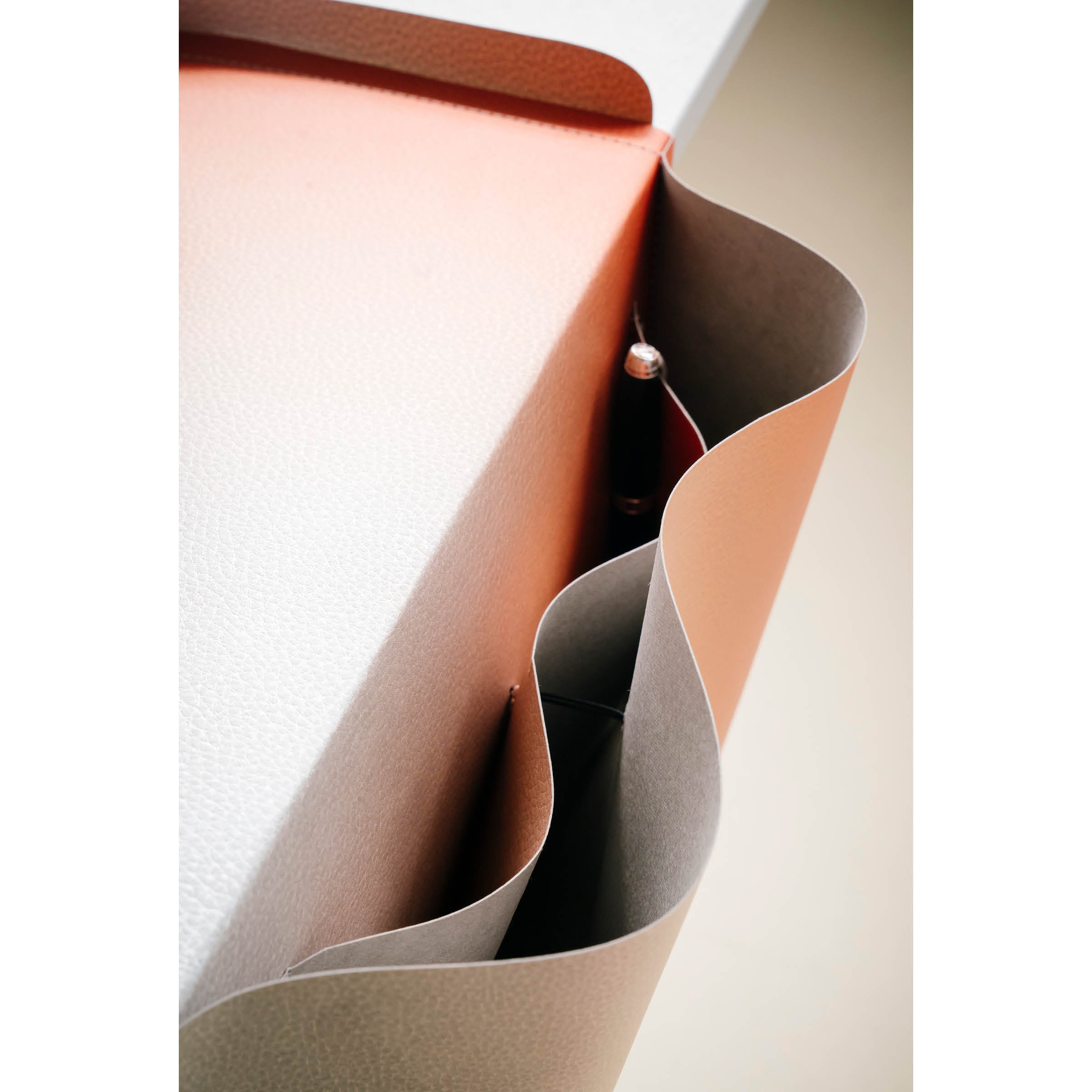 A close-up of the Salto Tray by Richard Lampert showcases a modern wavy-edged storage pocket made from sustainable Papyr® in Germany. It holds a pen and features a sleek, minimalist design with three-tone textured materials in orange, gray, and cream, attached to a table.