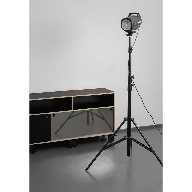 In a minimalistic room, an Egal Rockboard by Moormann, with its sleek black and white sliding doors and reflective glass, is positioned against a light gray floor and white wall. It captures the glossy shine of a spotlight on a tall tripod stand.