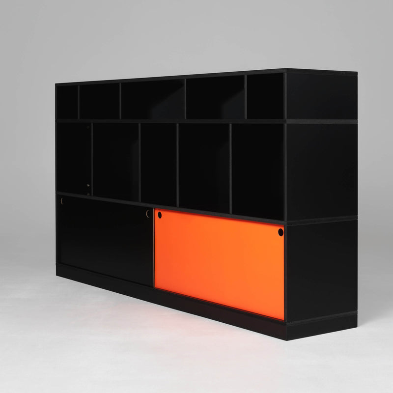 The Egal Highboard by Moormann is a black modular shelving unit featuring various-sized compartments and a bright orange steel sliding door, set against a neutral gray background.