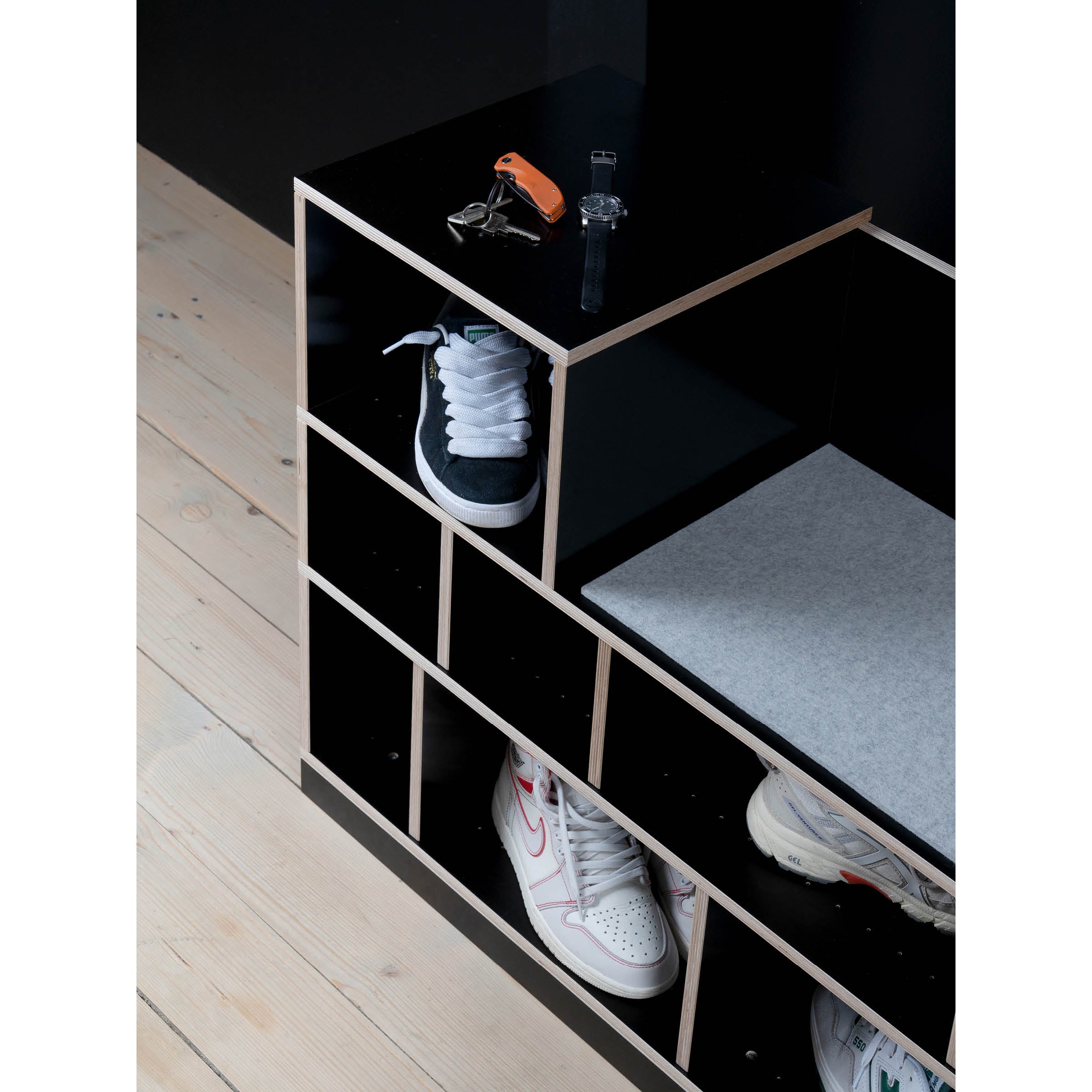 On a wooden floor, the Egal Sitzboard by Moormann, resembling a sleek sideboard, holds sneakers in its compartments. A cushion graces one side, while keys and a digital watch rest on top. Its all set against a dark wall backdrop.