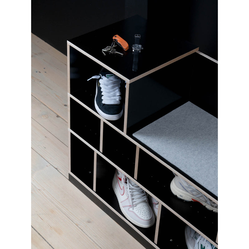 On a wooden floor, the Egal Sitzboard by Moormann, resembling a sleek sideboard, holds sneakers in its compartments. A cushion graces one side, while keys and a digital watch rest on top. Its all set against a dark wall backdrop.