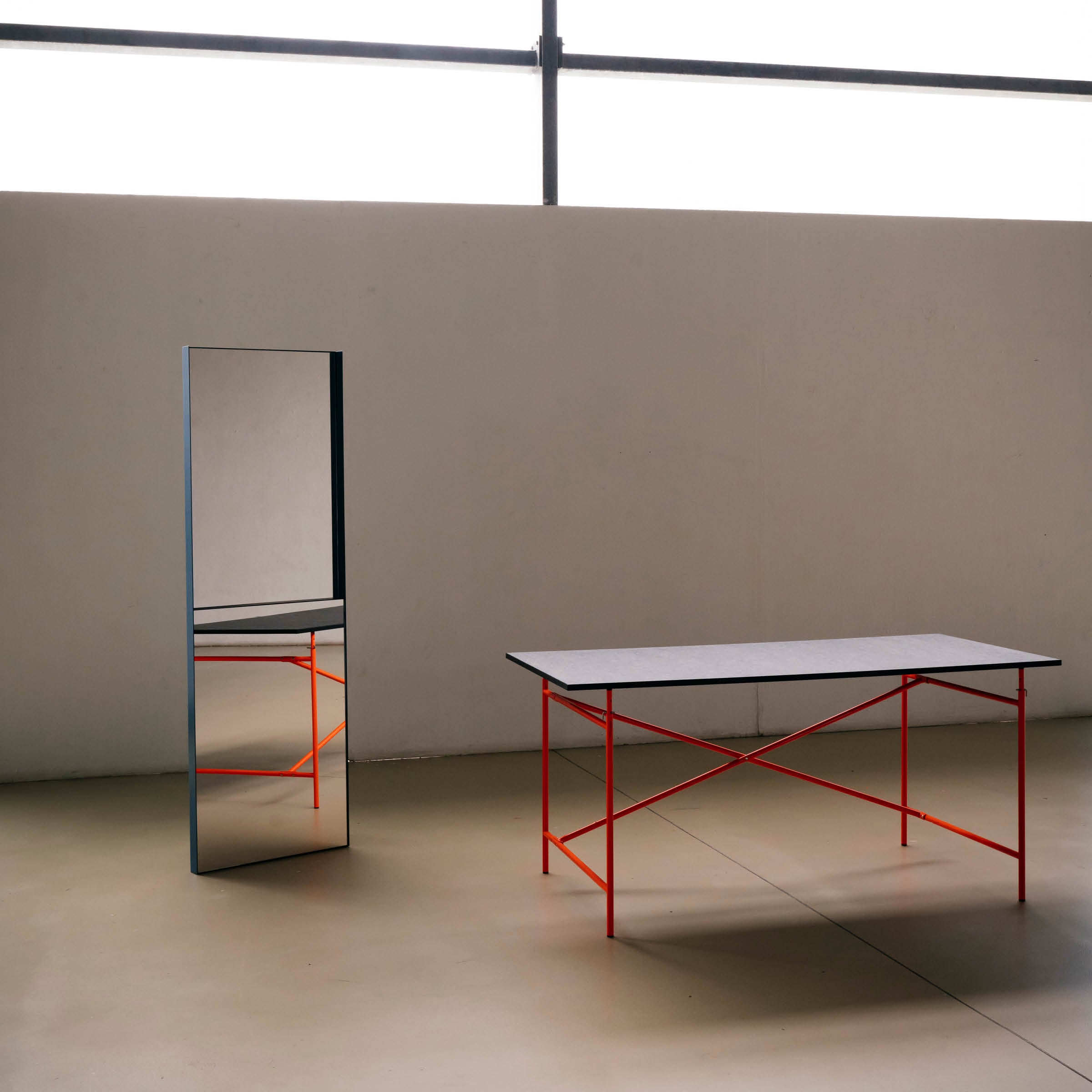 A minimalist room includes the OneTwo Mirror by Richard Lampert, reflecting a table with a black top and bright red legs on a beige floor. The light wall features horizontal windows above, ideal for contemporary home accessory enthusiasts.