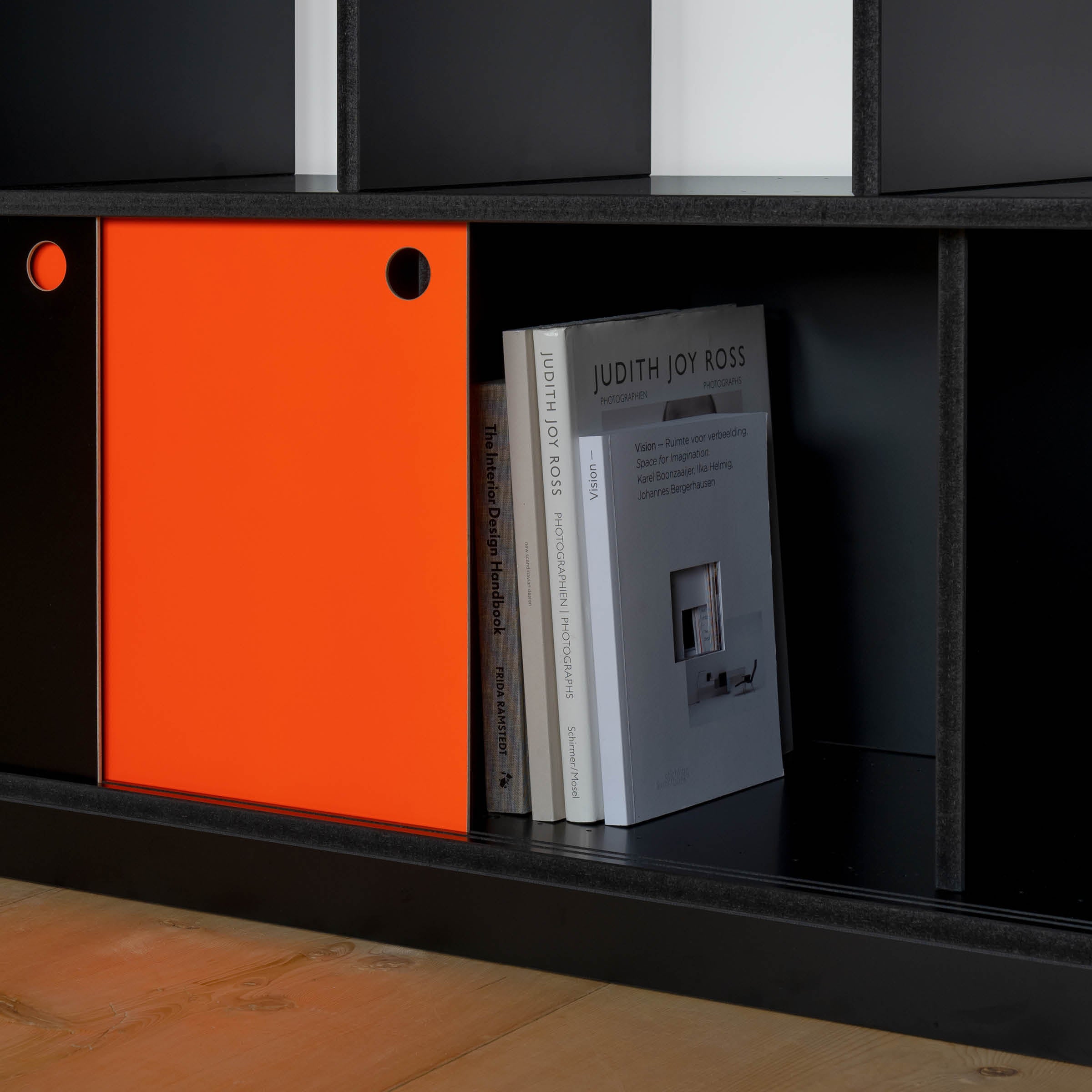 The Egal Highboard by Moormann, in sleek black with a vibrant orange sliding door, reveals three upright books. Its elegant design and steel accents create a stunning contrast against the light wooden floor.