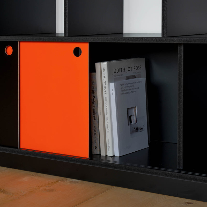 The Egal Highboard by Moormann, in sleek black with a vibrant orange sliding door, reveals three upright books. Its elegant design and steel accents create a stunning contrast against the light wooden floor.