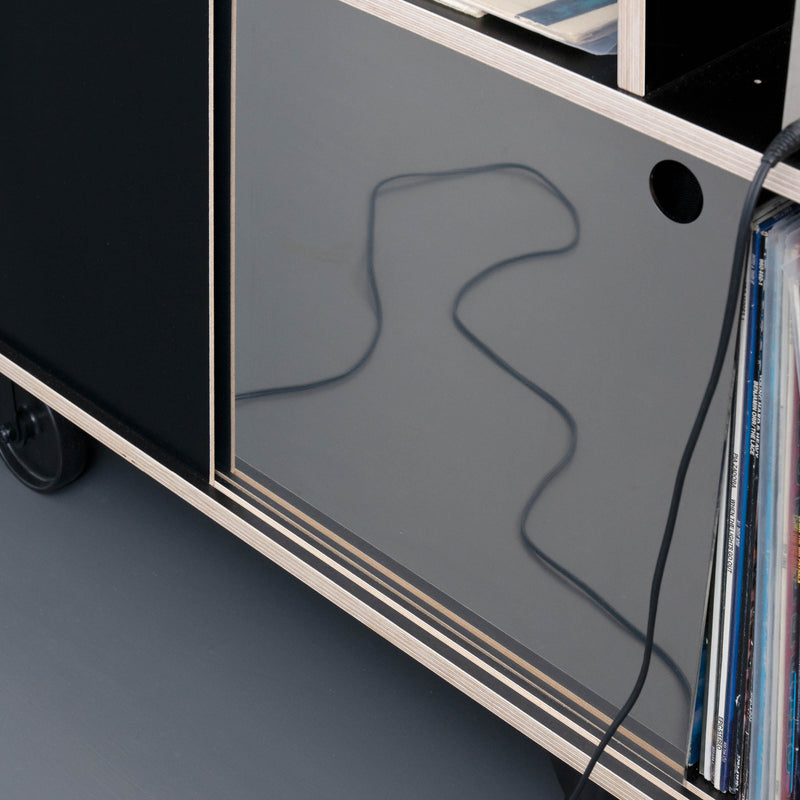 The Egal Rockboard by Moormann is a contemporary sideboard with glossy black panels and wooden edges. Stainless steel wheels support it on a gray floor, while shelves partially visible house vinyl records, and a cord casts shadows over its sleek surface.