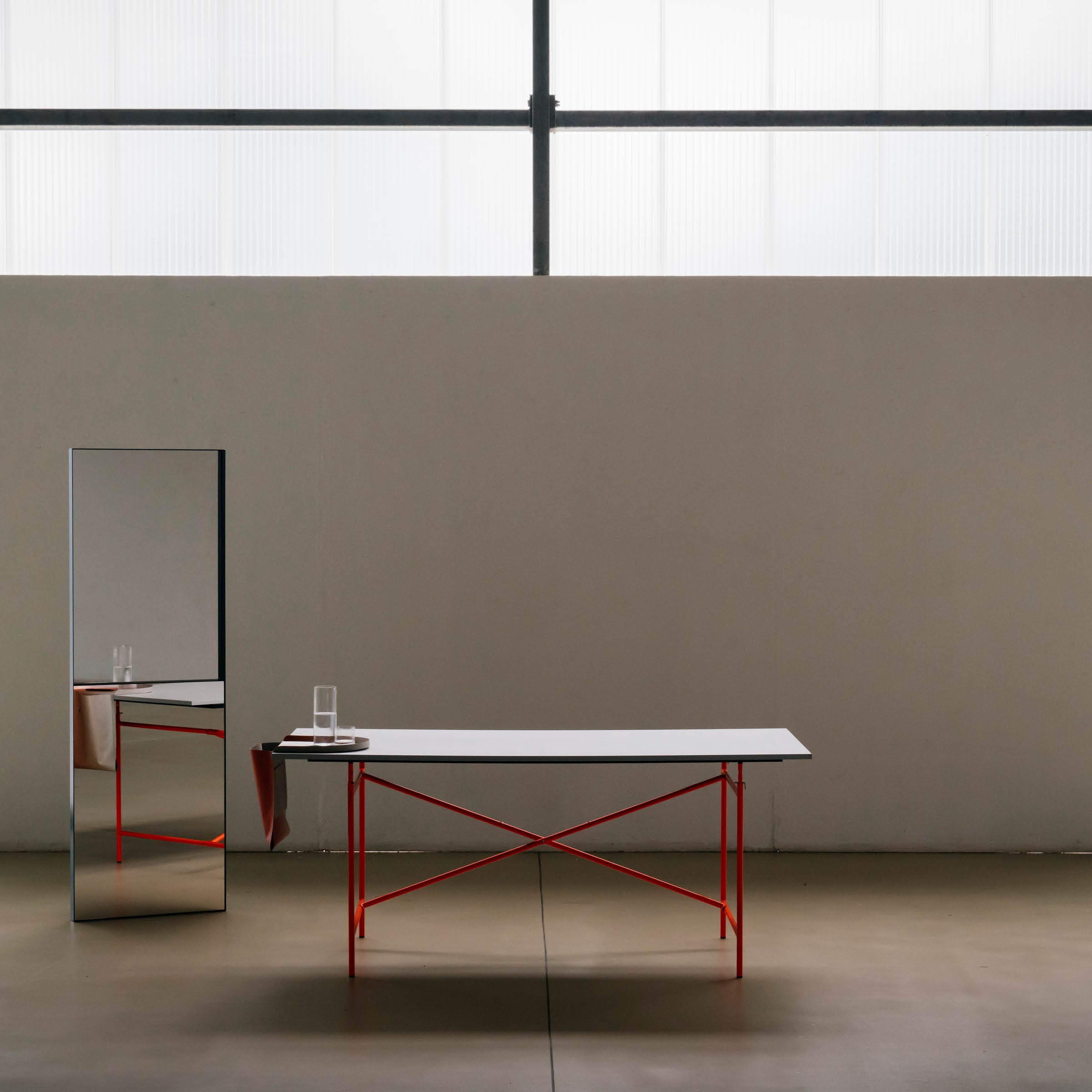 A minimalist room features a Richard Lampert Salto Tray with a white Papyr® tabletop and red legs. A tall, narrow mirror reflects the table, which holds books and a glass of water. Gray walls and floor enhance the natural light from the large window above. Made sustainably in Germany.