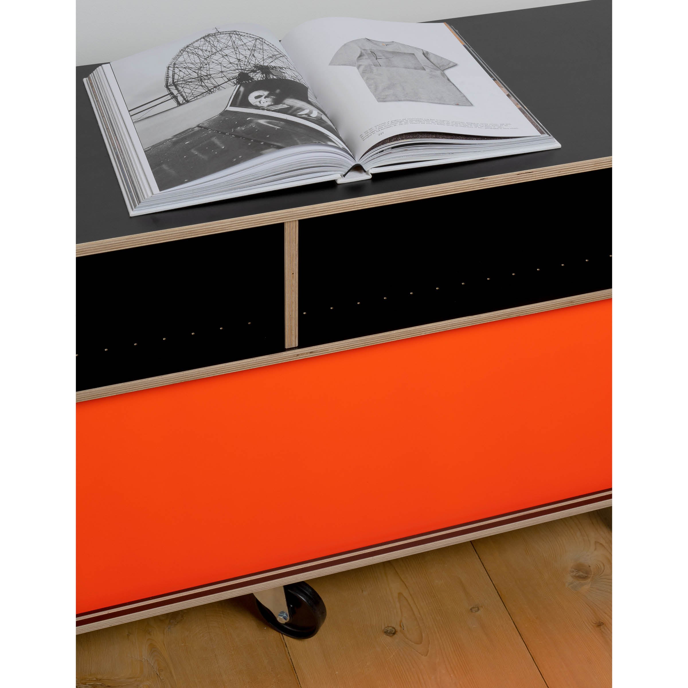 An open book sits on a Moormann Egal Rollboard, showcasing its sleek black and red design with wood accents and wheels. The light brown wood floor enhances the modern look, while sliding doors subtly frame black and white images inside, including one of a shirt.