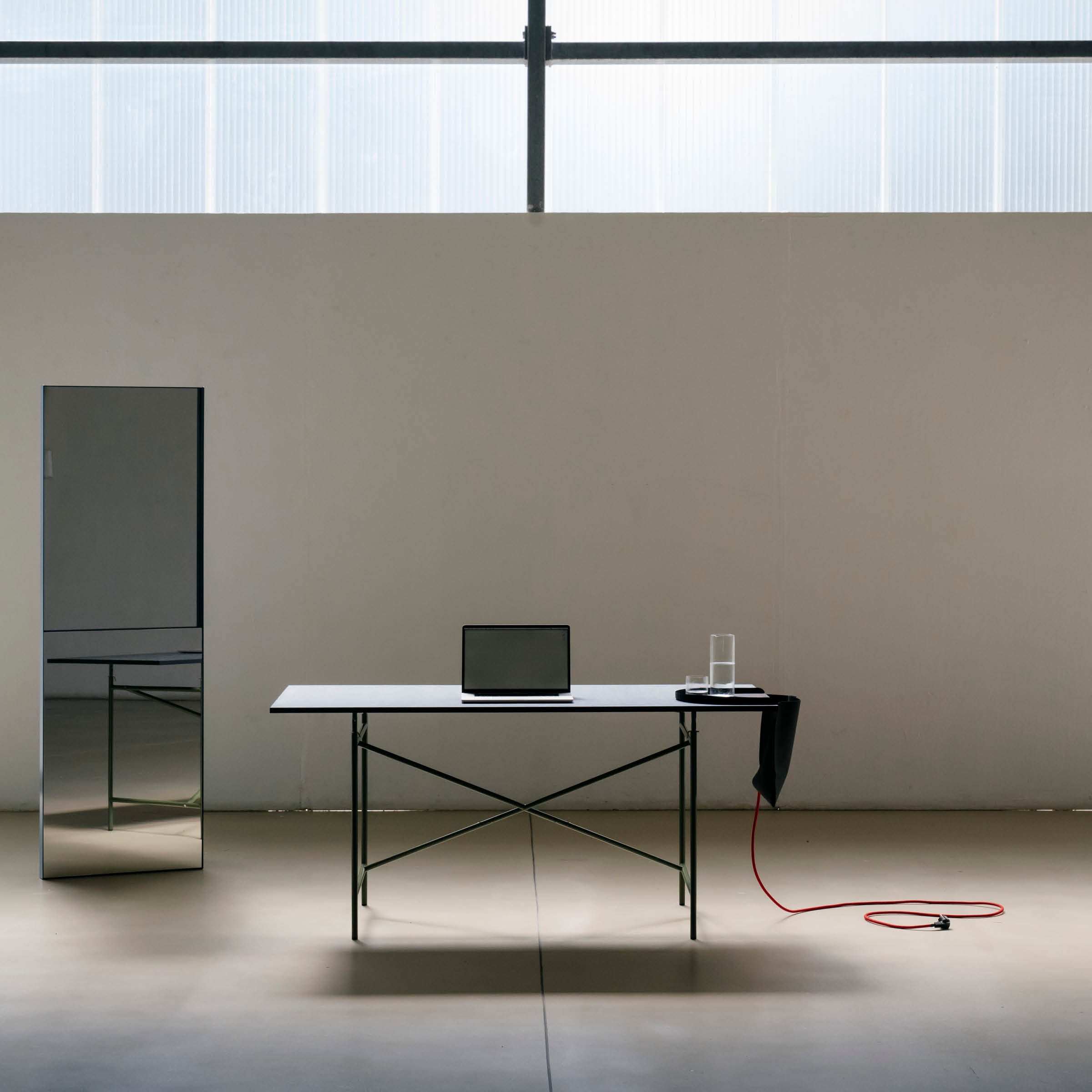 In a minimalist room, a Salto Tray by Richard Lampert centers the table with a closed laptop and water glass. A black Papyr® material cloth drapes the edge, while a red cable lays on the floor. A tall mirror on the left reflects light from a large window, embodying sustainable elegance perfectly.
