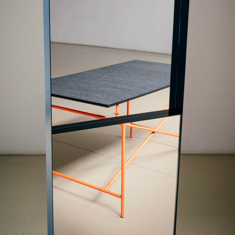 In a minimalist space, the OneTwo Mirror by Richard Lampert reflects a modern table with a dark top and sleek orange legs against beige flooring—a perfect setup for contemporary home accessory enthusiasts.