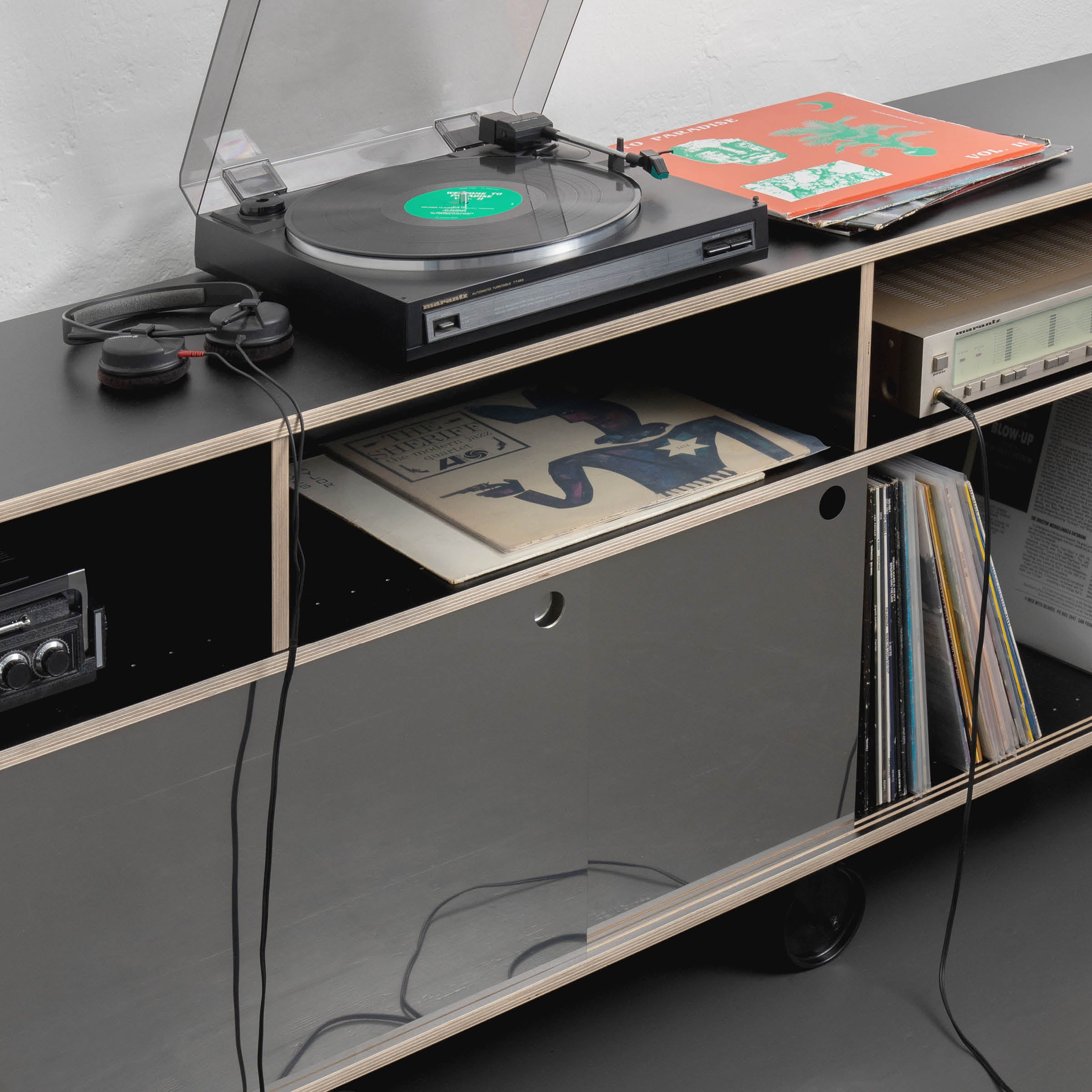 The Egal Rockboard by Moormann, a sleek black media console, features sliding doors concealing audio equipment and vinyl records. The setup includes a record player and headphones, with nearby record sleeves and subtle stainless steel accents, creating a retro minimalist vibe.