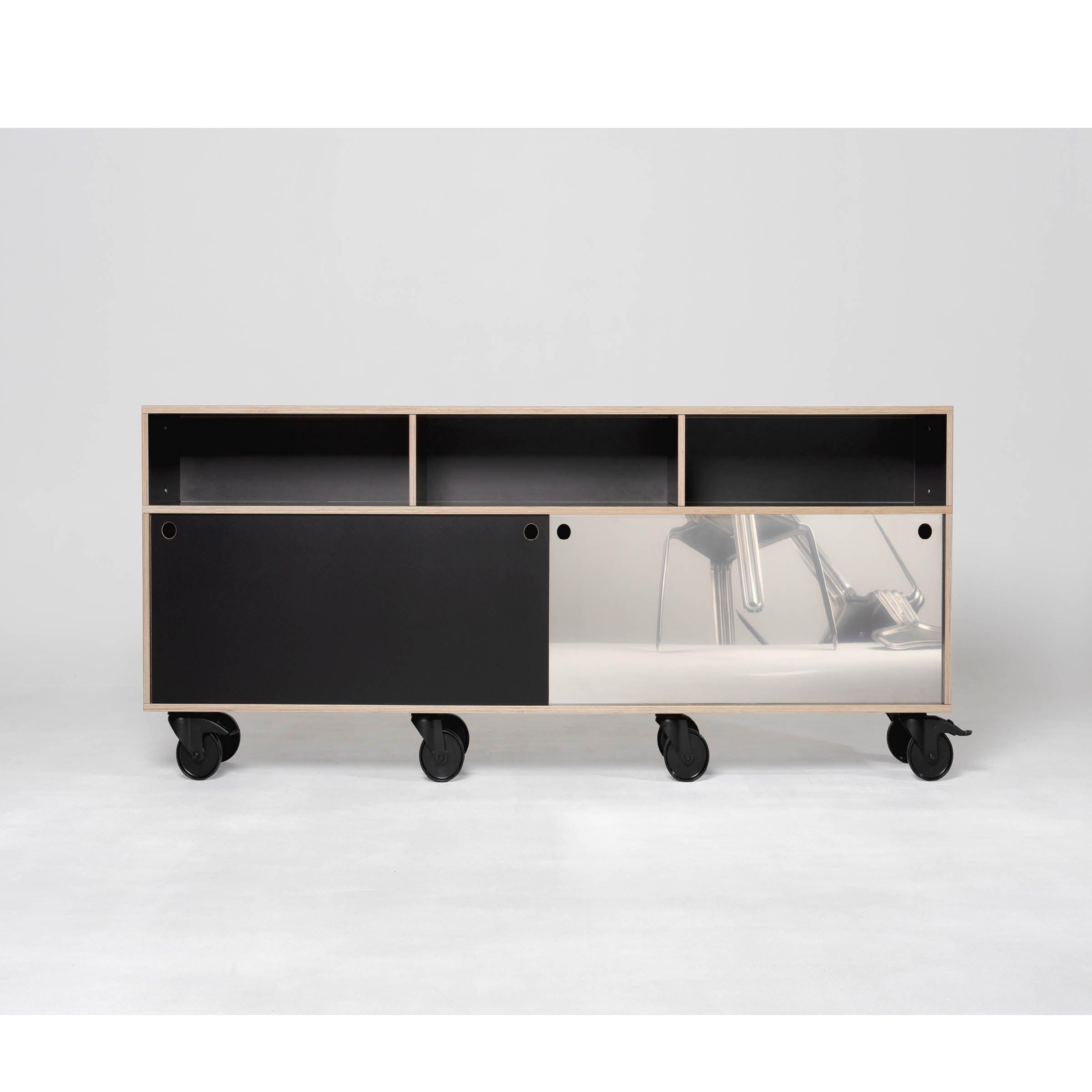 The Egal Rockboard by Moormann is a minimalist mobile sideboard featuring black and white design, open shelves, a sliding door, and wheels. A partly clear bottom panel reveals a stainless steel object inside, set against a plain white background.