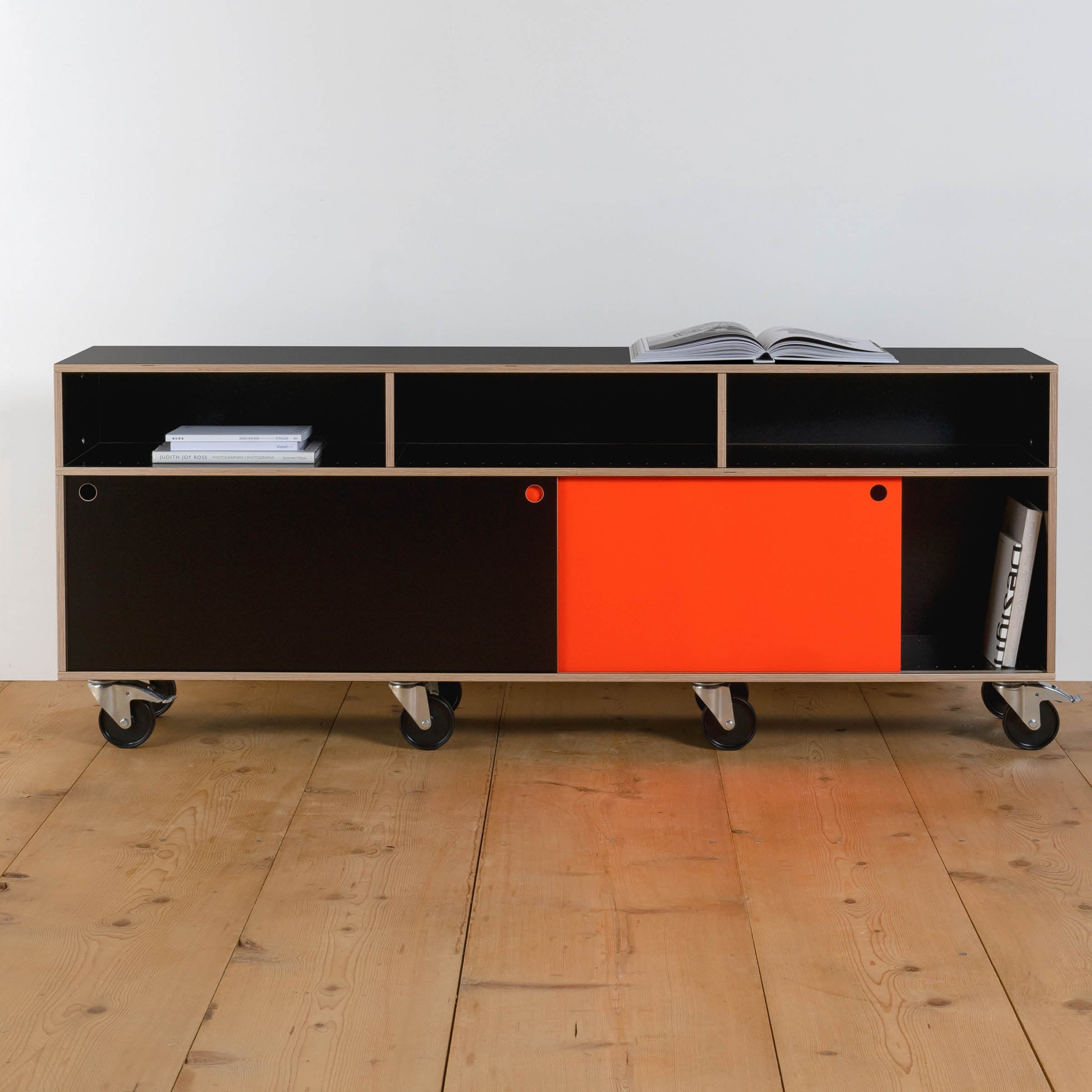 The Moormann Egal Rollboard, a modern minimalist rolling cabinet with sleek black and red sliding doors, graces the wooden floor. It features open shelves for displaying books and magazines, while four caster wheels offer effortless mobility.