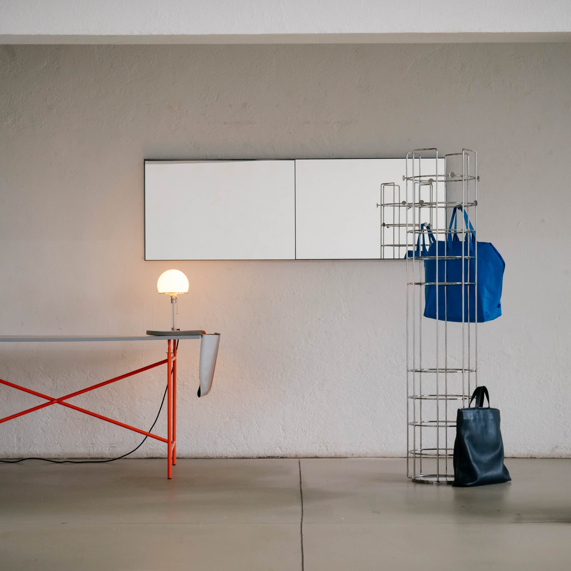 A minimalist interior showcases Richard Lamperts OneTwo Mirror on the wall, a metal storage rack with two blue bags, and a low table with a red frame. Concrete flooring and plain walls maintain simplicity.