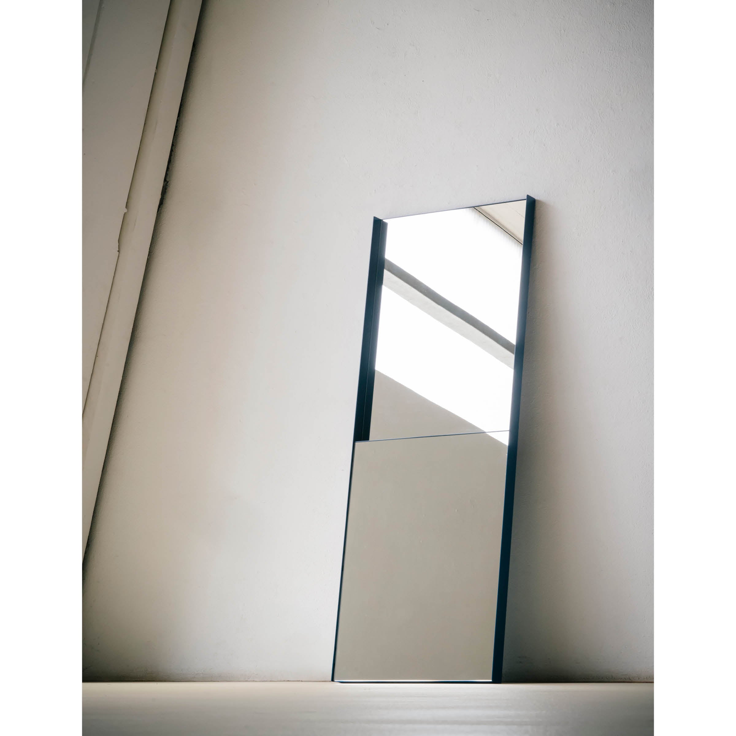 The OneTwo Mirror by Richard Lampert, a tall rectangular piece with a minimalist metal frame, leans stylishly against a white wall. Its sleek design reflects the rooms ceiling and beige wall, enhancing its contemporary charm, while the light floor complements the settings minimalism.