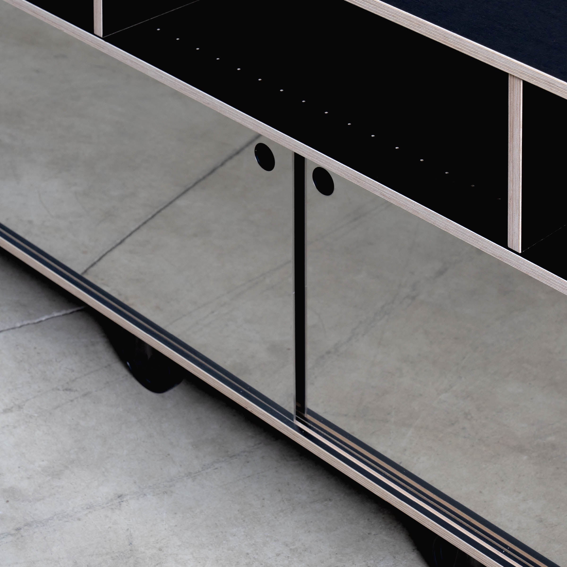 A close-up of the Egal Rockboard by Moormann showcases its modern design, featuring reflective glass sliding doors with black circular handles, mounted on wheels. The sleek style includes light wood edges and stainless steel accents against a concrete floor background.
