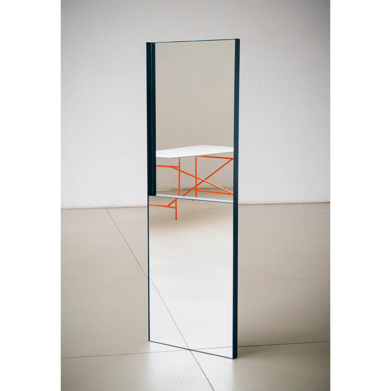 The OneTwo Mirror by Richard Lampert is a tall, narrow freestanding piece with a minimalist metal frame. It reflects part of a room featuring a white table with an orange base, enhancing the clean, modern aesthetic against light-colored floors and walls.
