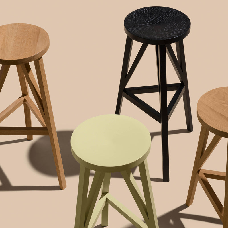 The JL4 Faber Barstools by Loehr feature four uniquely silhouetted wooden stools against a light beige backdrop. They come in light brown, black, beige, and medium brown, complementing their solid oak build. Each stool has three legs with triangular bracing for support, casting shadows below.