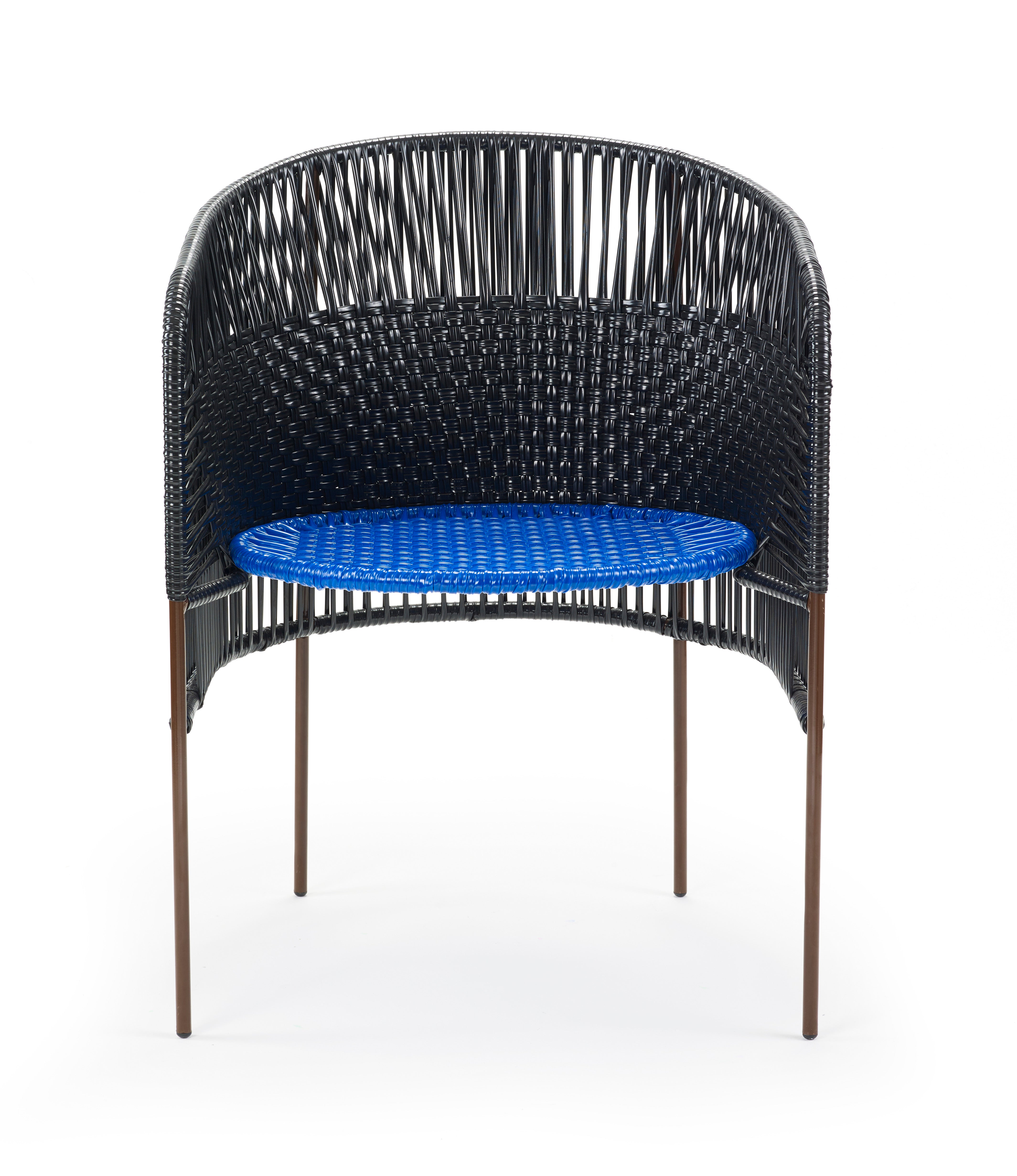 The Caribe Dining Chair by Ames features a curved backrest and woven black details with a bright blue seat. Inspired by Colombian weaving, it has slim brown metal legs and an open design, perfect for chic outdoor settings.