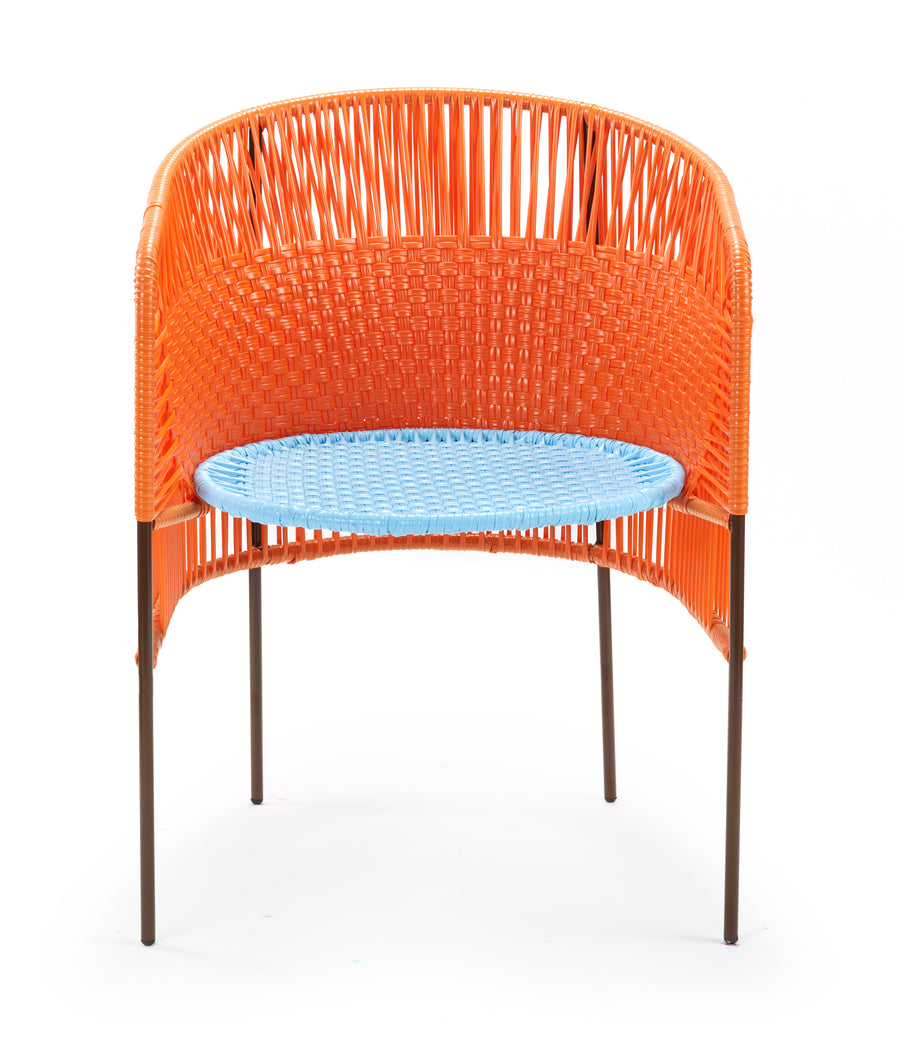 The Ames Caribe Dining Chair features Colombian weaving in orange on the back and sides, a round blue woven seat, and slim brown metal legs—perfect as standout outdoor furniture against a white background.