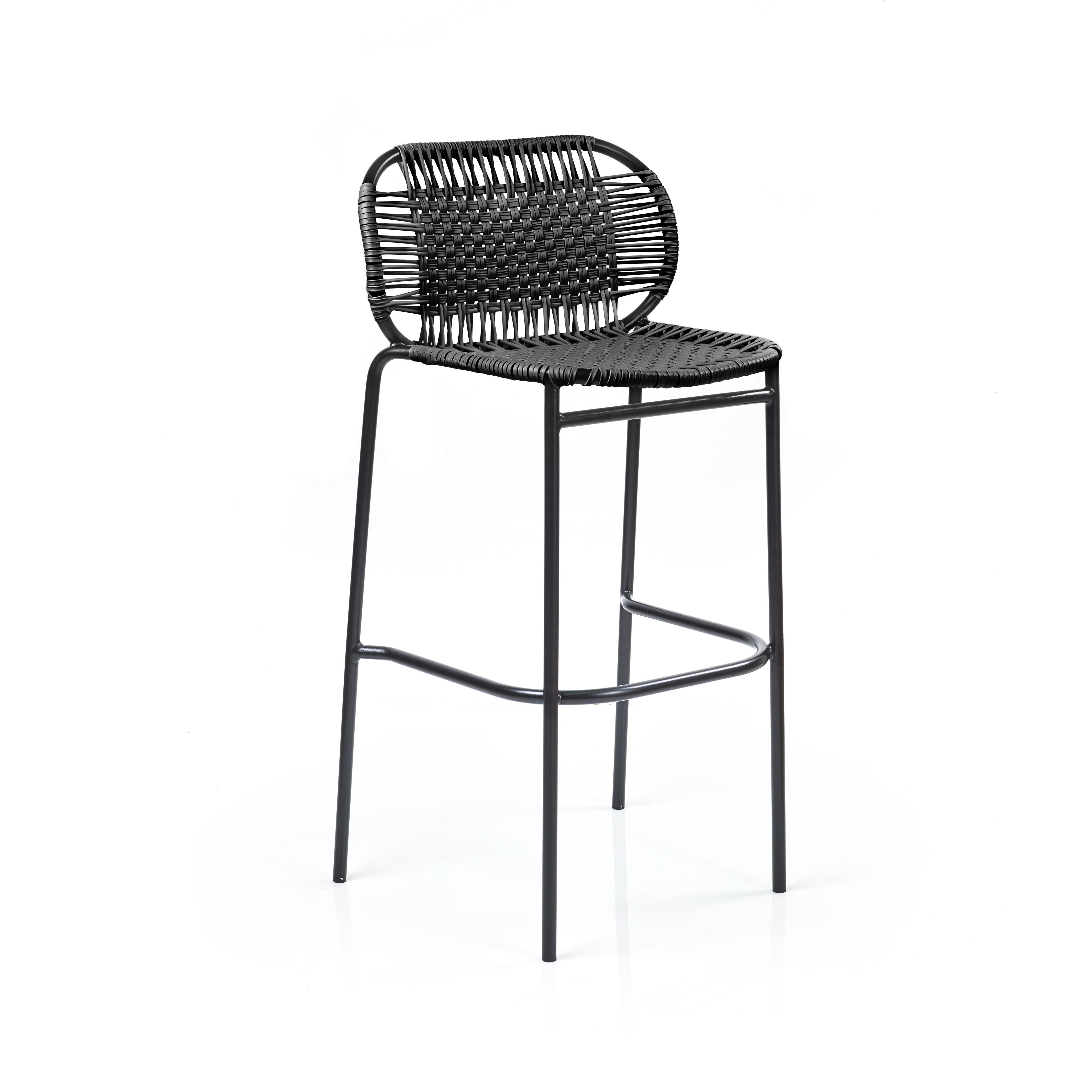 The Ames Cielo Barstool by Ames features a black, woven rattan-style backrest and seat on slim metal legs with a footrest, offering a sleek, modern look reminiscent of Sebastian Herkner’s style against a plain white background.