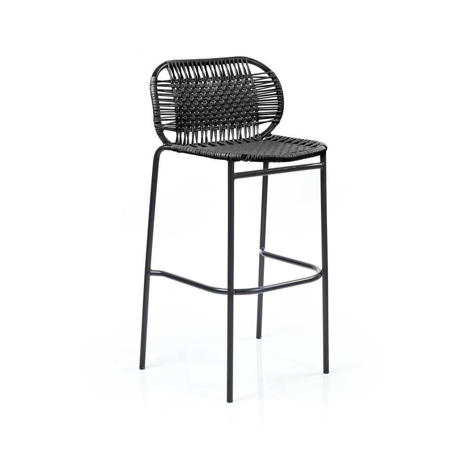 The Ames Cielo Barstool by Ames features a black, woven rattan-style backrest and seat on slim metal legs with a footrest, offering a sleek, modern look reminiscent of Sebastian Herkner’s style against a plain white background.