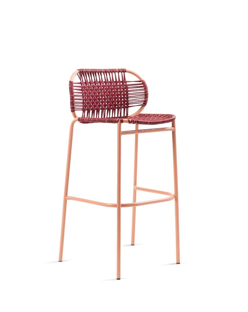 The Ames Cielo Barstool by Ames, designed by Sebastian Herkner, features a peach metal frame, red and pink woven seat and backrest with a rounded back, footrest, and stackable design for modern practicality.