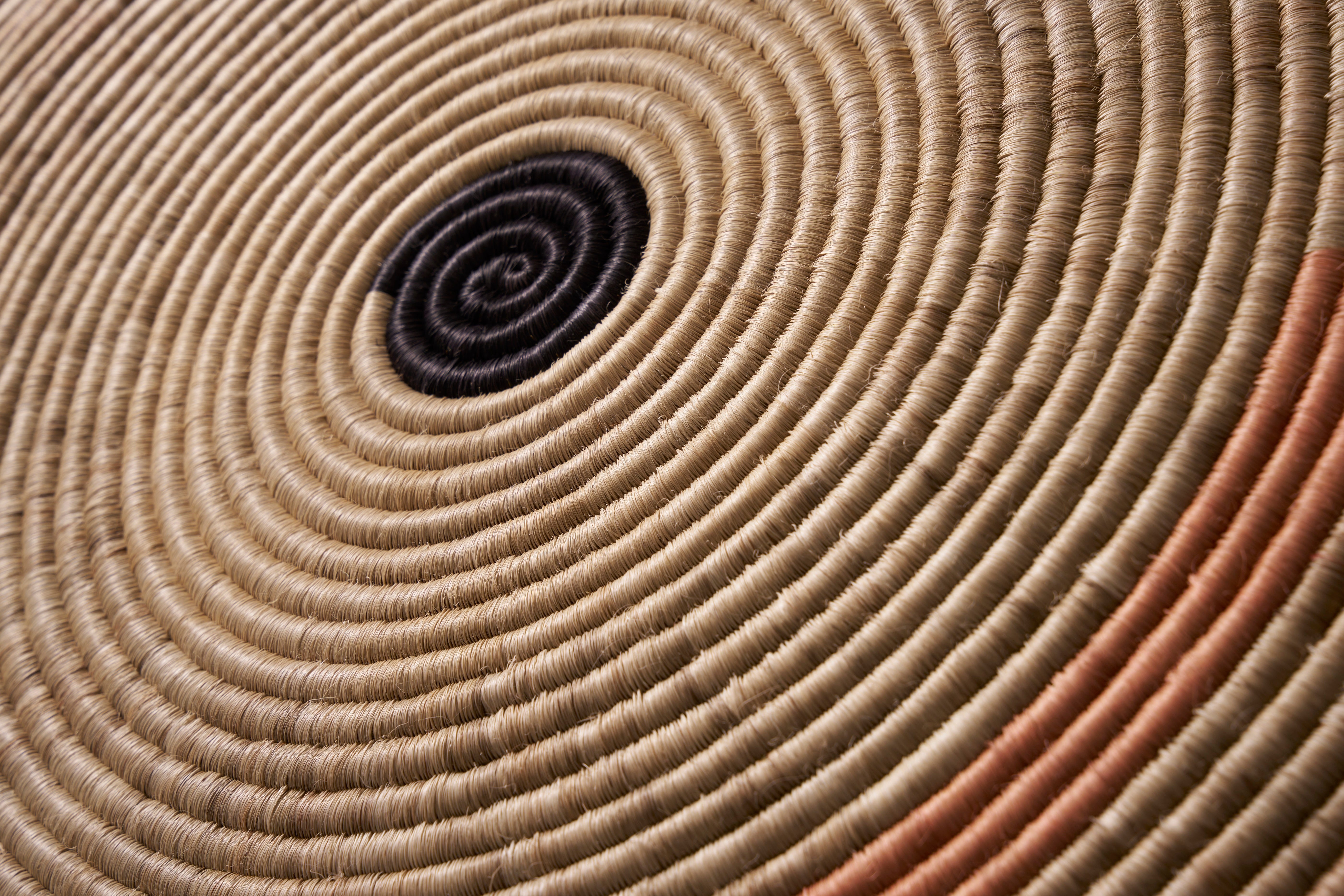 Close-up of the Ames Ojitos Wall Decoration Andes by Ames, a woven basket with tightly coiled fique palm fibers in concentric circles, featuring a black center with subtle tan and orange hues—ideal as wall décor.