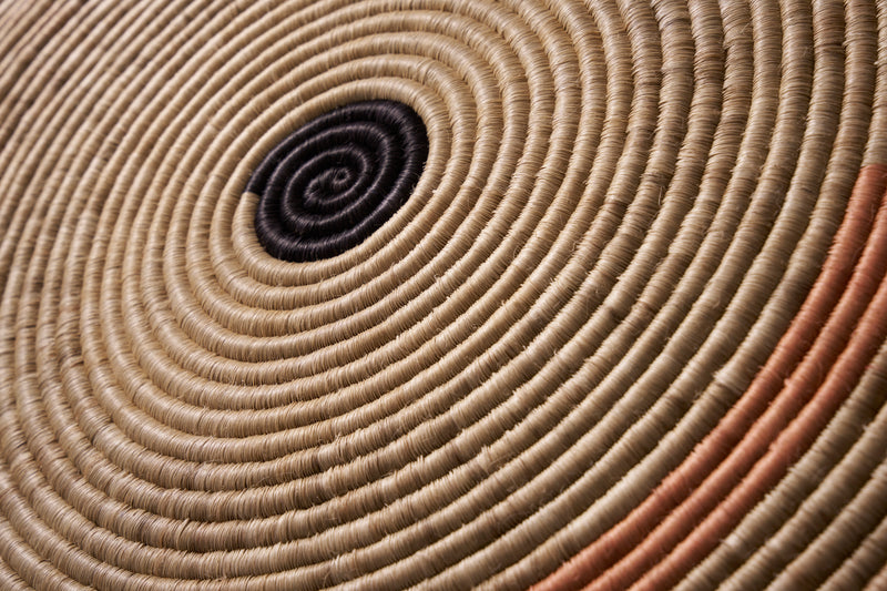 Close-up of the Ames Ojitos Wall Decoration Andes by Ames, a woven basket with tightly coiled fique palm fibers in concentric circles, featuring a black center with subtle tan and orange hues—ideal as wall décor.