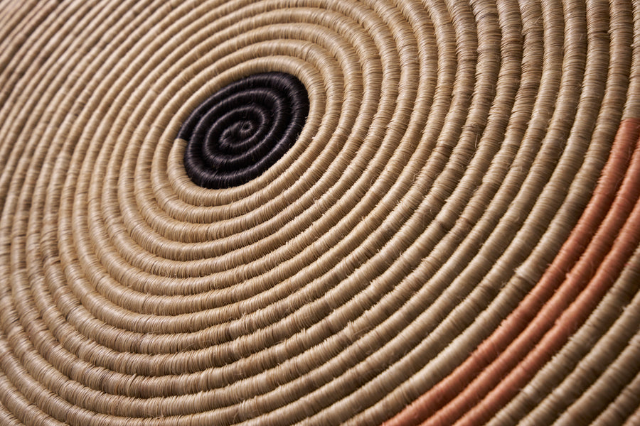 Close-up of the Ames Ojitos Wall Decoration Andes by Ames, a woven basket with tightly coiled fique palm fibers in concentric circles, featuring a black center with subtle tan and orange hues—ideal as wall décor.
