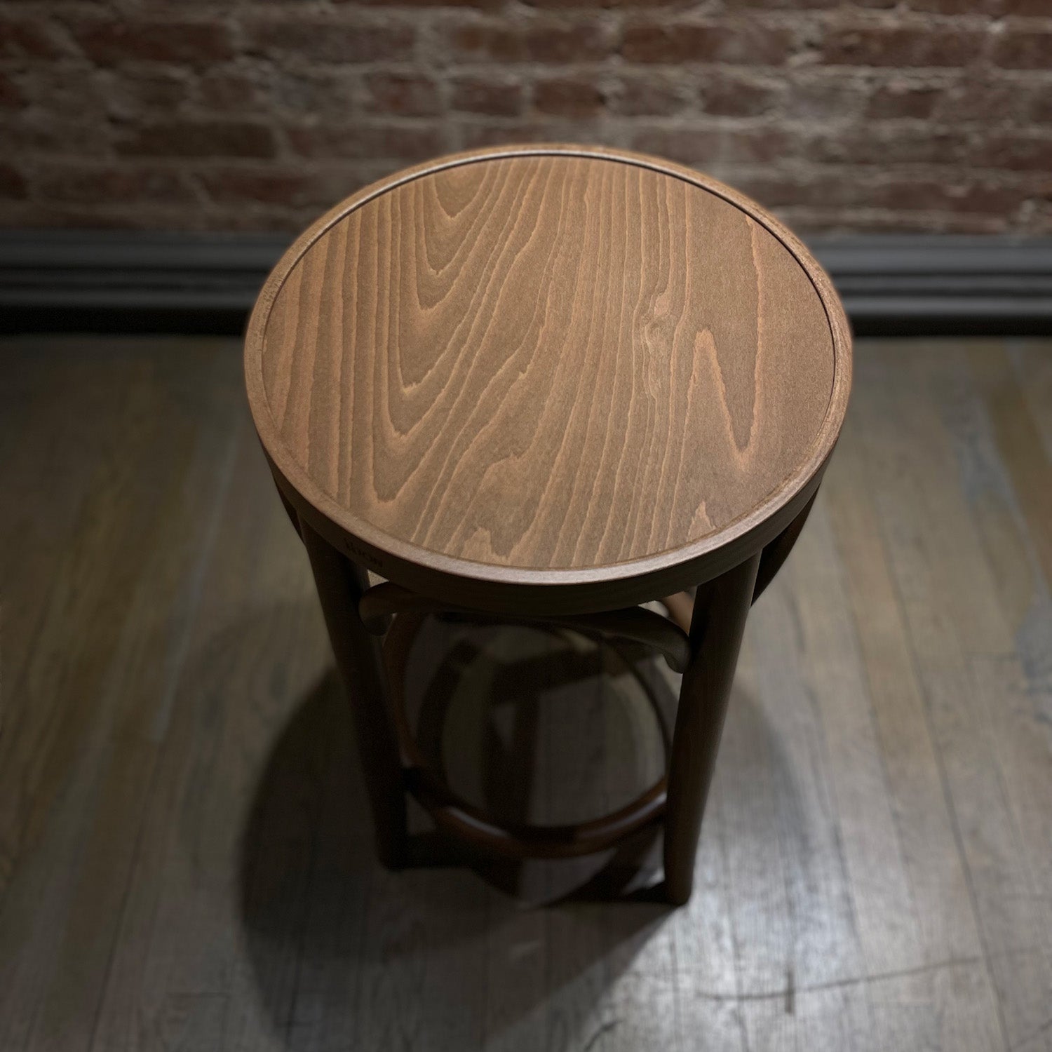 A Barstool 73 by TON, featuring a small, round wooden design with beech leg joints and a smooth top, sits on a hardwood floor. The brick wall background and lighting create a soft shadow around the stool, highlighting its decorative base.