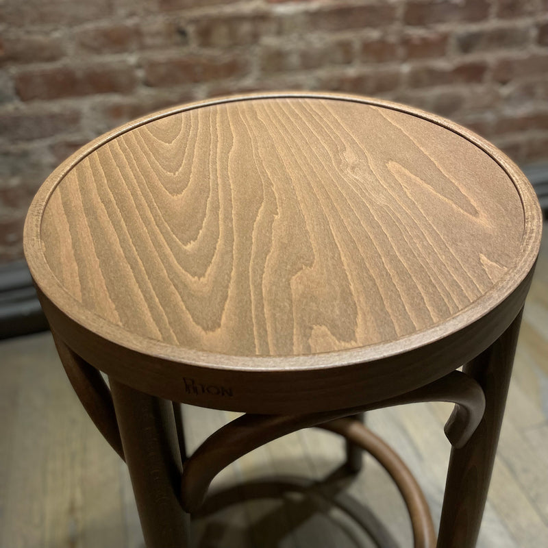 A close-up of the Barstool 73 by TON features a round wooden seat with natural grain and beech leg joints. Its rustic appearance is set against a wooden floor and brick wall background, enhanced by an upholstered seat for comfort, with THONET subtly engraved on the side.