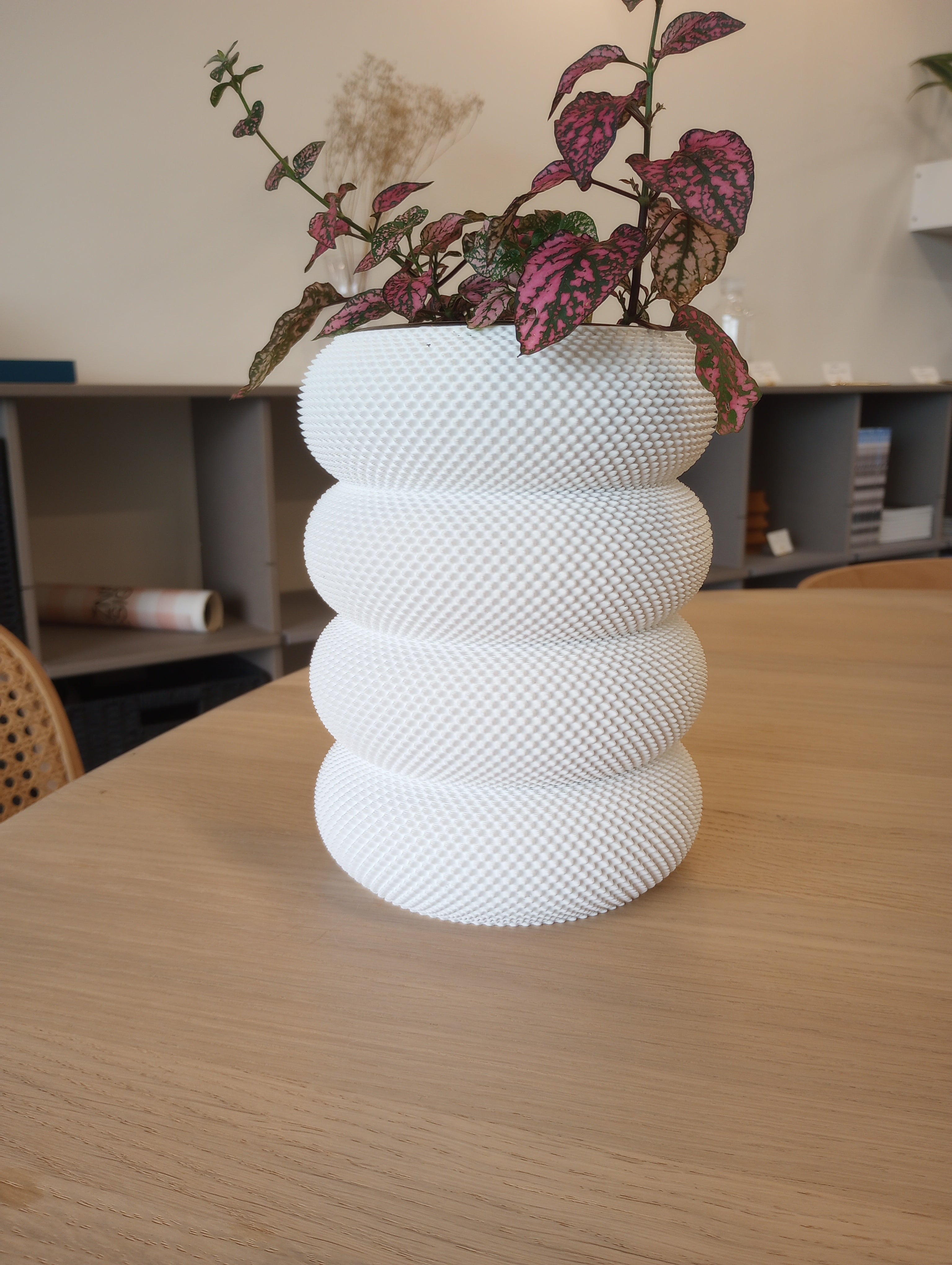 The Dorn 341g 1c Vase by Sheyn, a white textured vessel with a stacked ring design crafted eco-consciously, adorns a wooden table holding a pink and green-leafed plant. Shelves and furniture are visible in the background.